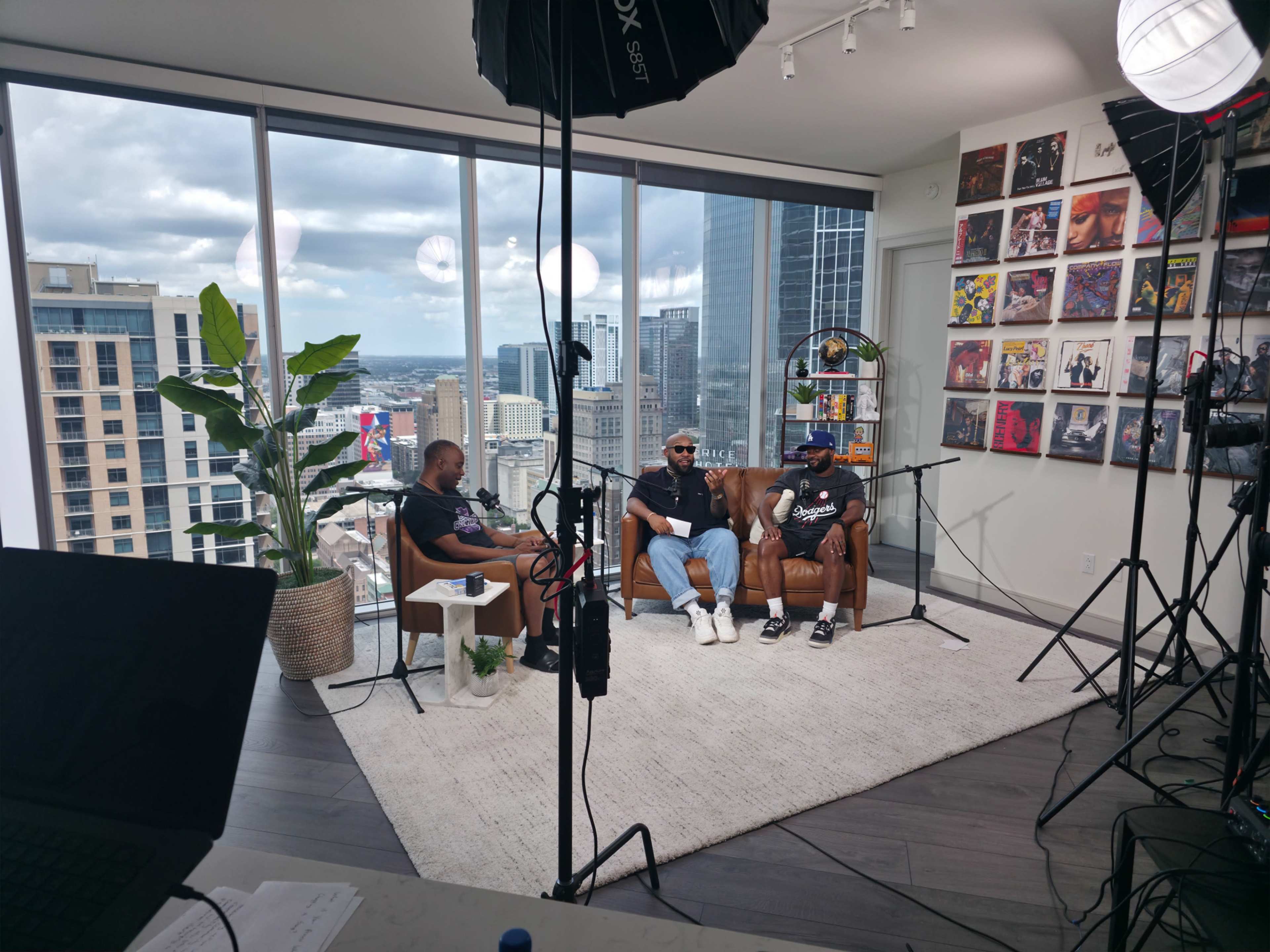 The image shows two people sitting on a sofa in a modern, well-lit studio with large windows overlooking a city skyline, surrounded by cameras and lighting equipment.
