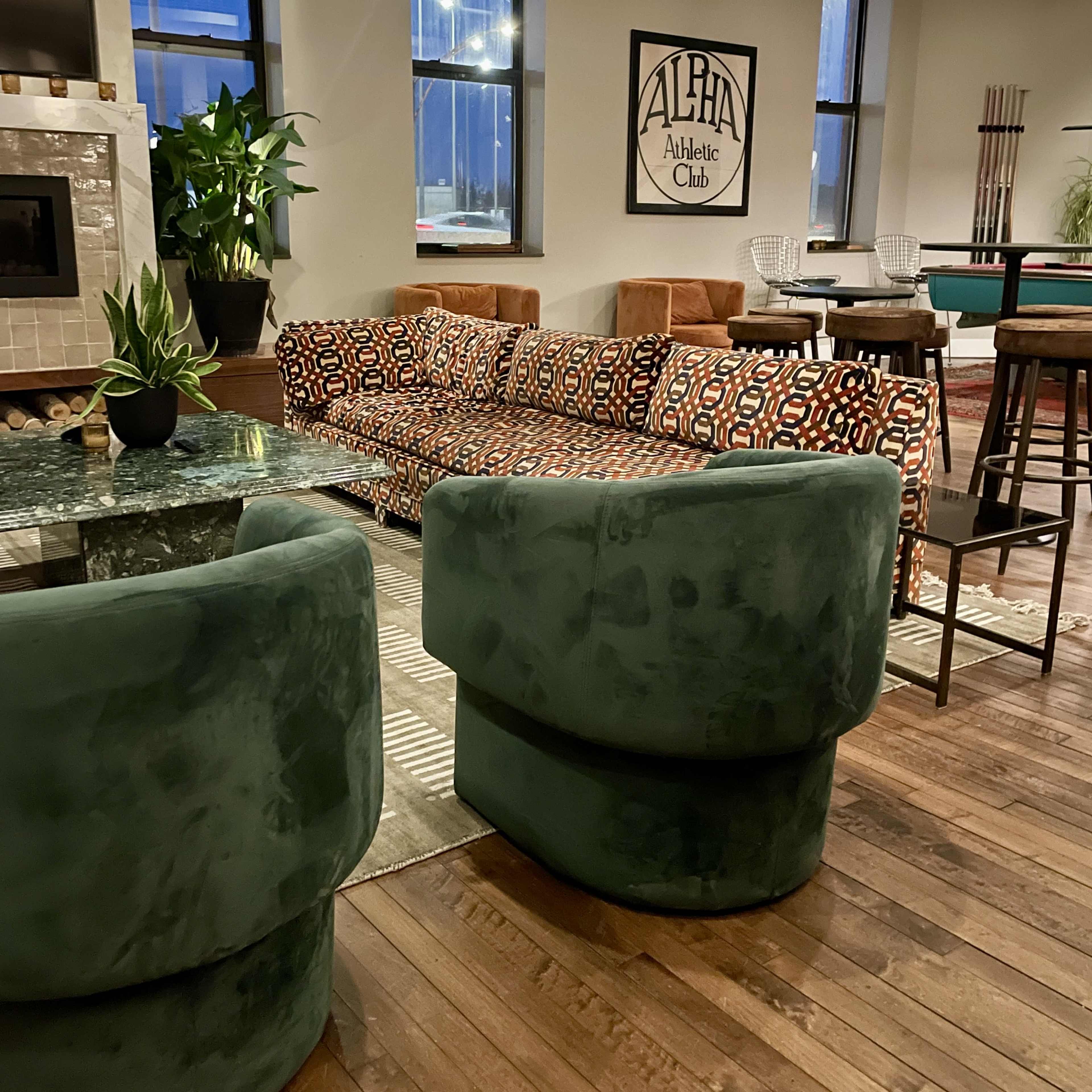 The image shows a modern lounge area with a patterned sofa, green velvet chairs, a glass table, and a wall featuring a sign for "Alpha Athletic Club."