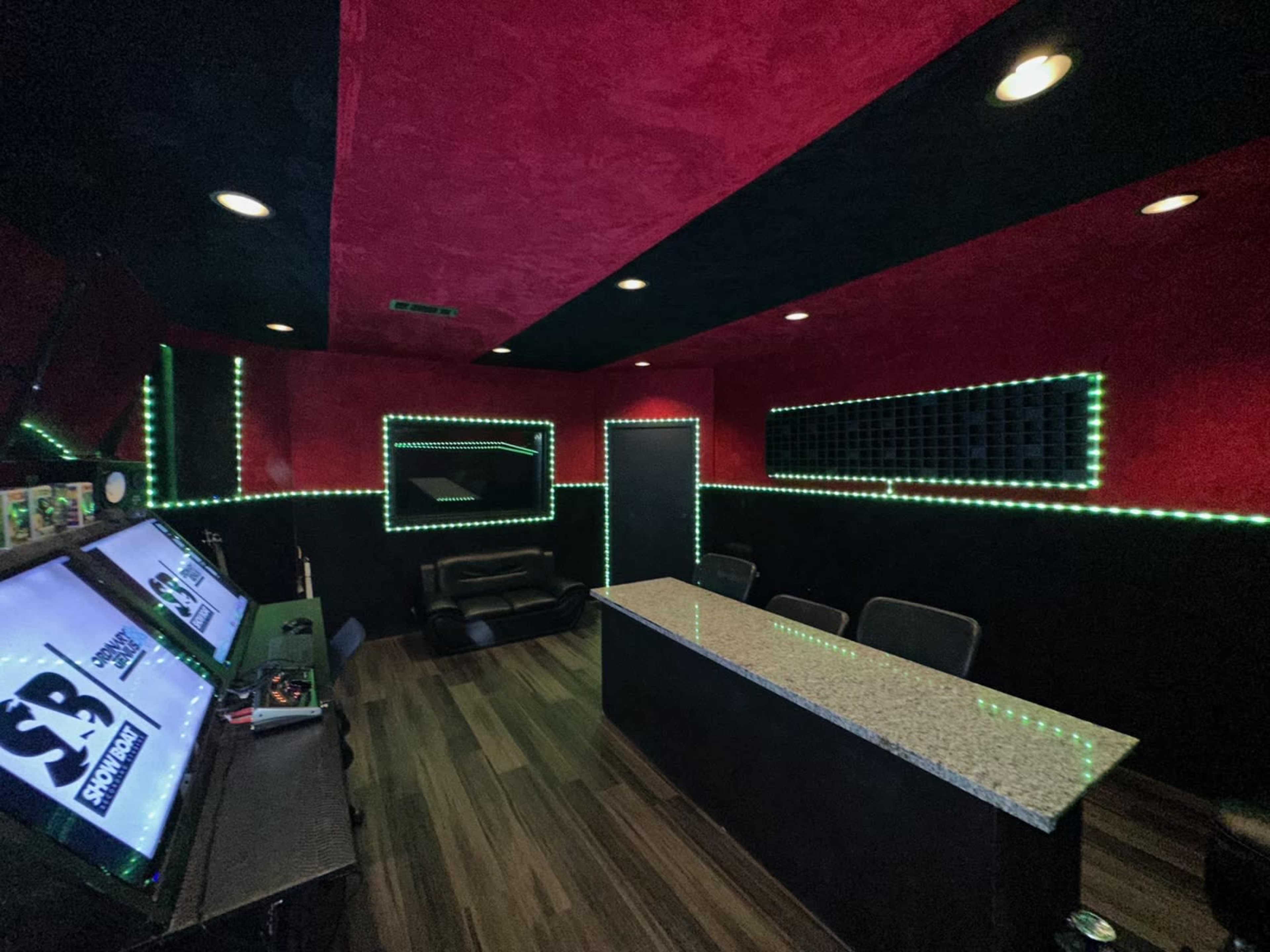 The image shows a dimly lit room with red and black walls, featuring a long counter, a black couch, and green LED lights along the walls and ceiling.