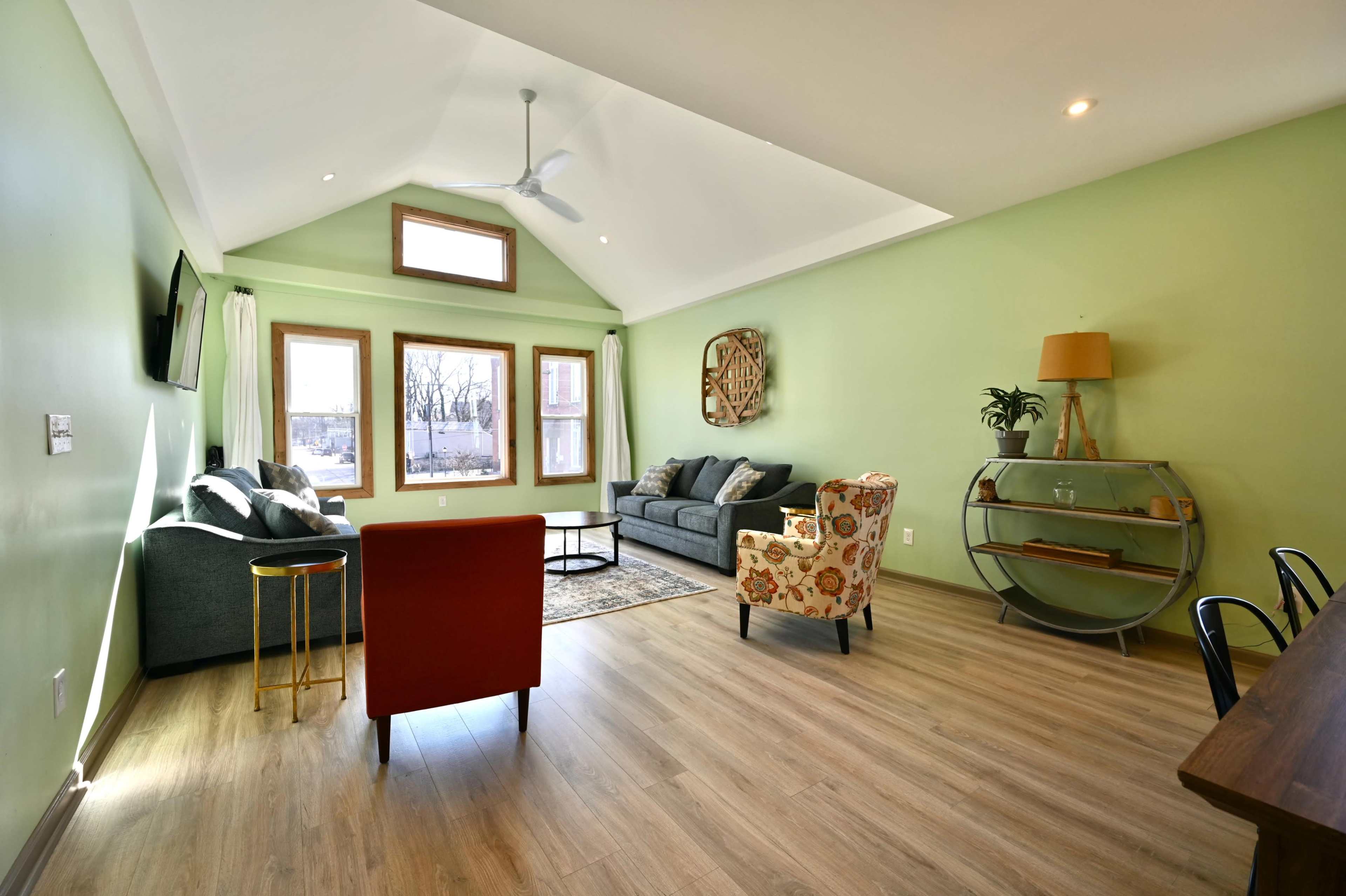 A contemporary living room with green walls, hardwood flooring, a gray sofa, an armchair with a floral pattern, and a glass coffee table.