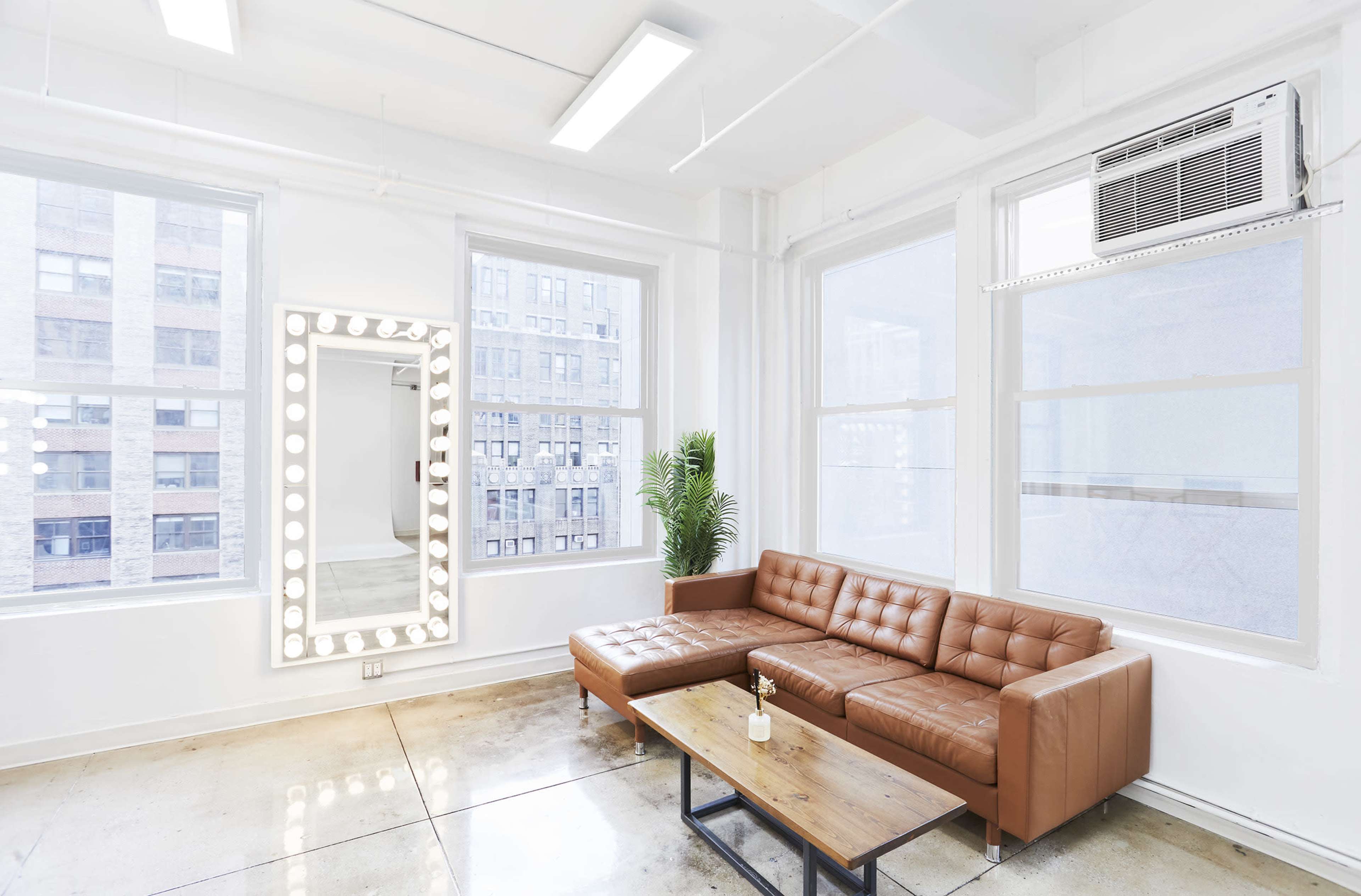 A modern lounge area features a brown leather sectional sofa, a wooden coffee table, a large illuminated mirror, and large windows allowing natural light.