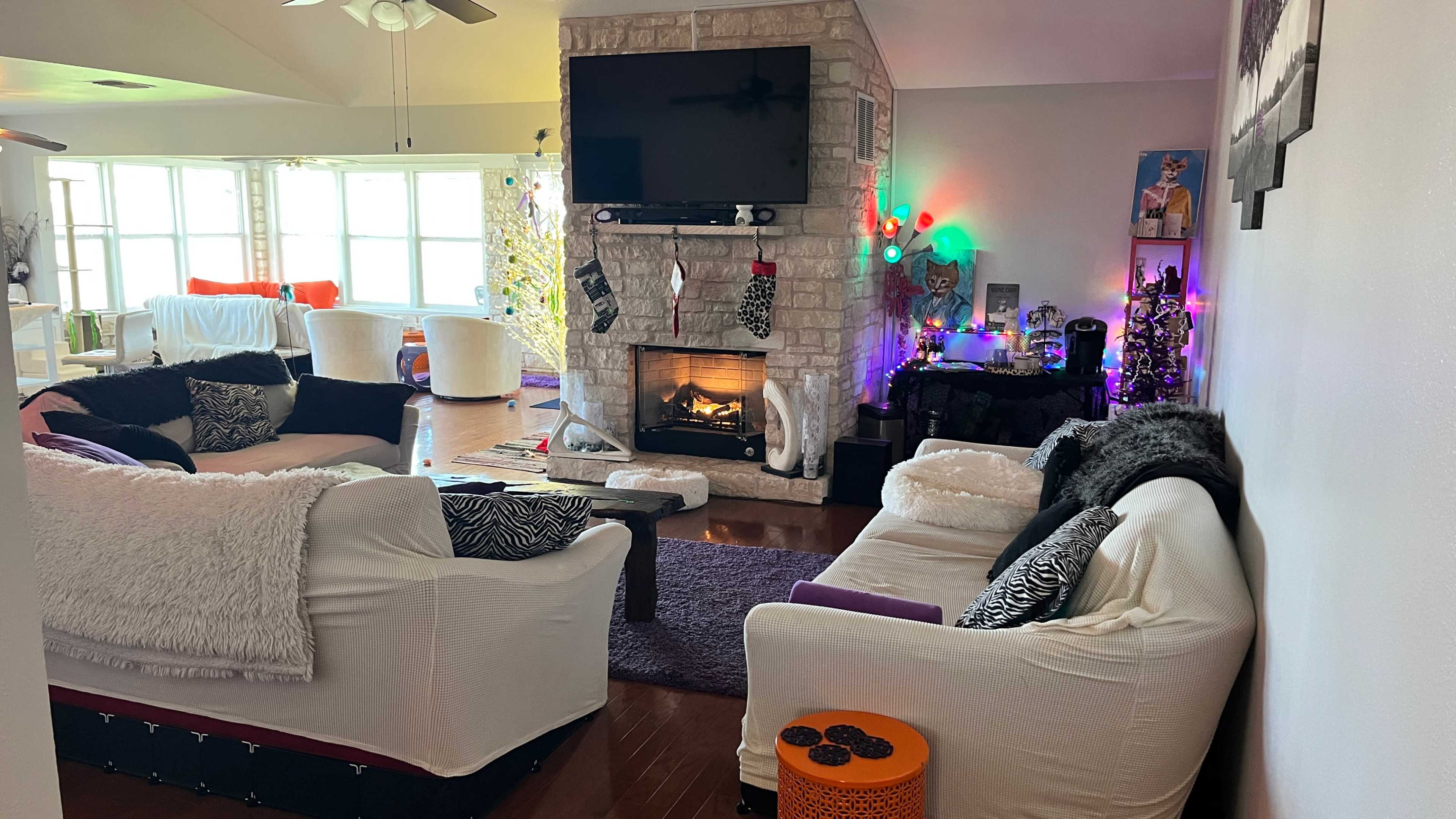A cozy living room features two sofas, a fireplace with stockings, and colorful holiday decorations.
