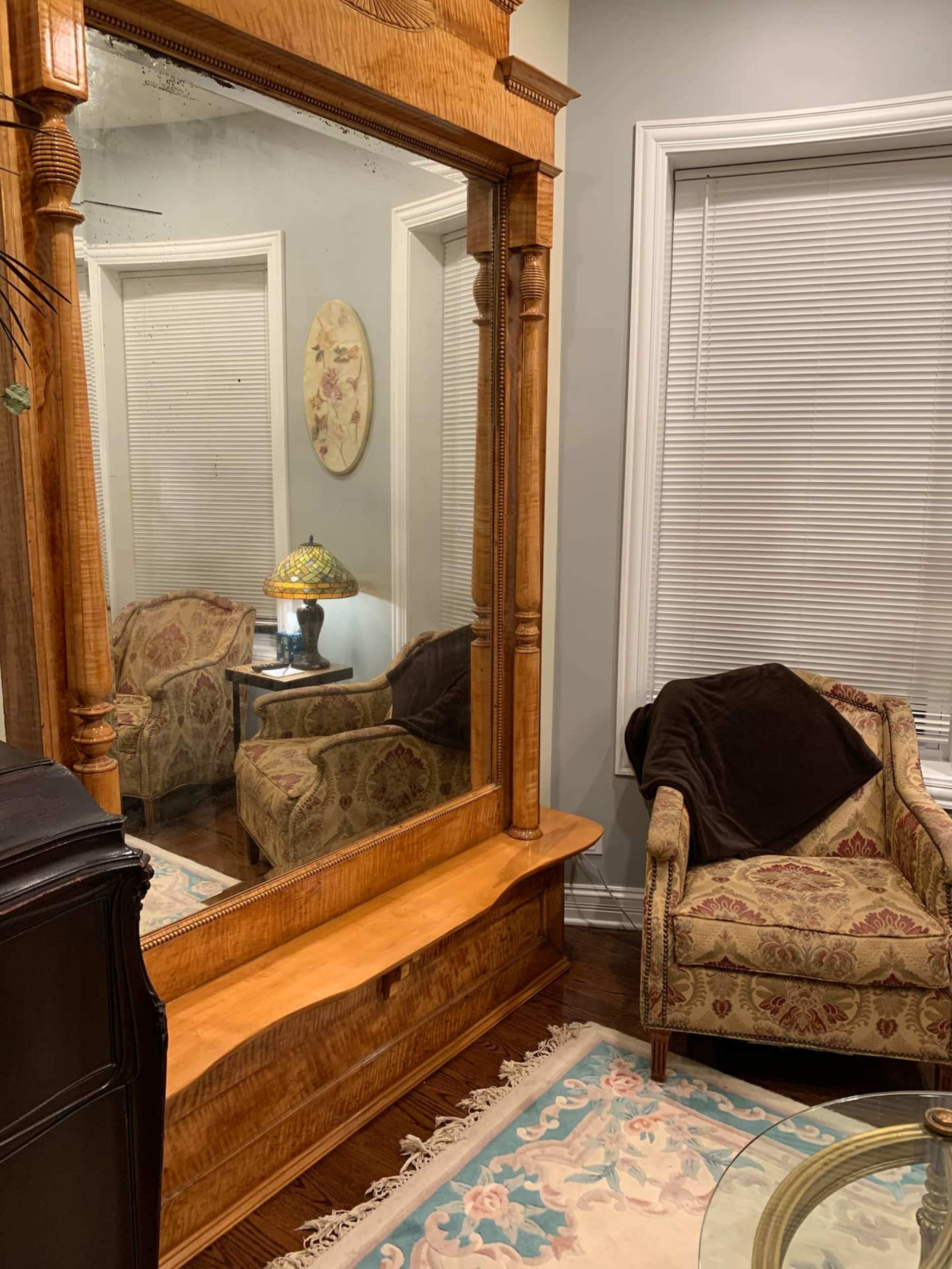 The image shows a large ornate wooden mirror reflecting a cozy corner with a patterned armchair and a lamp.