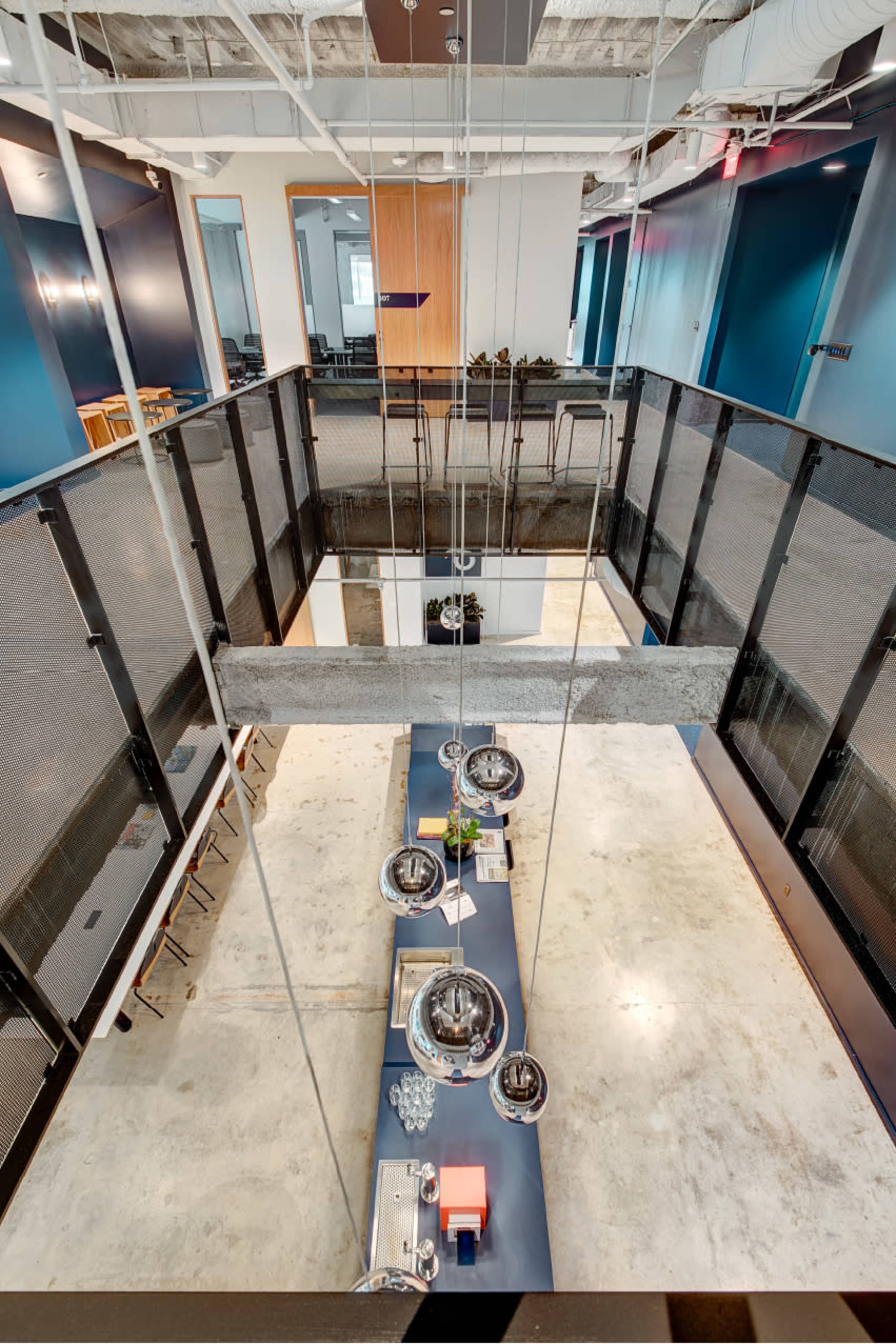 A modern open-plan office space with a sleek, suspended lighting fixture above a central meeting area, surrounded by glass partitions and seating arrangements.