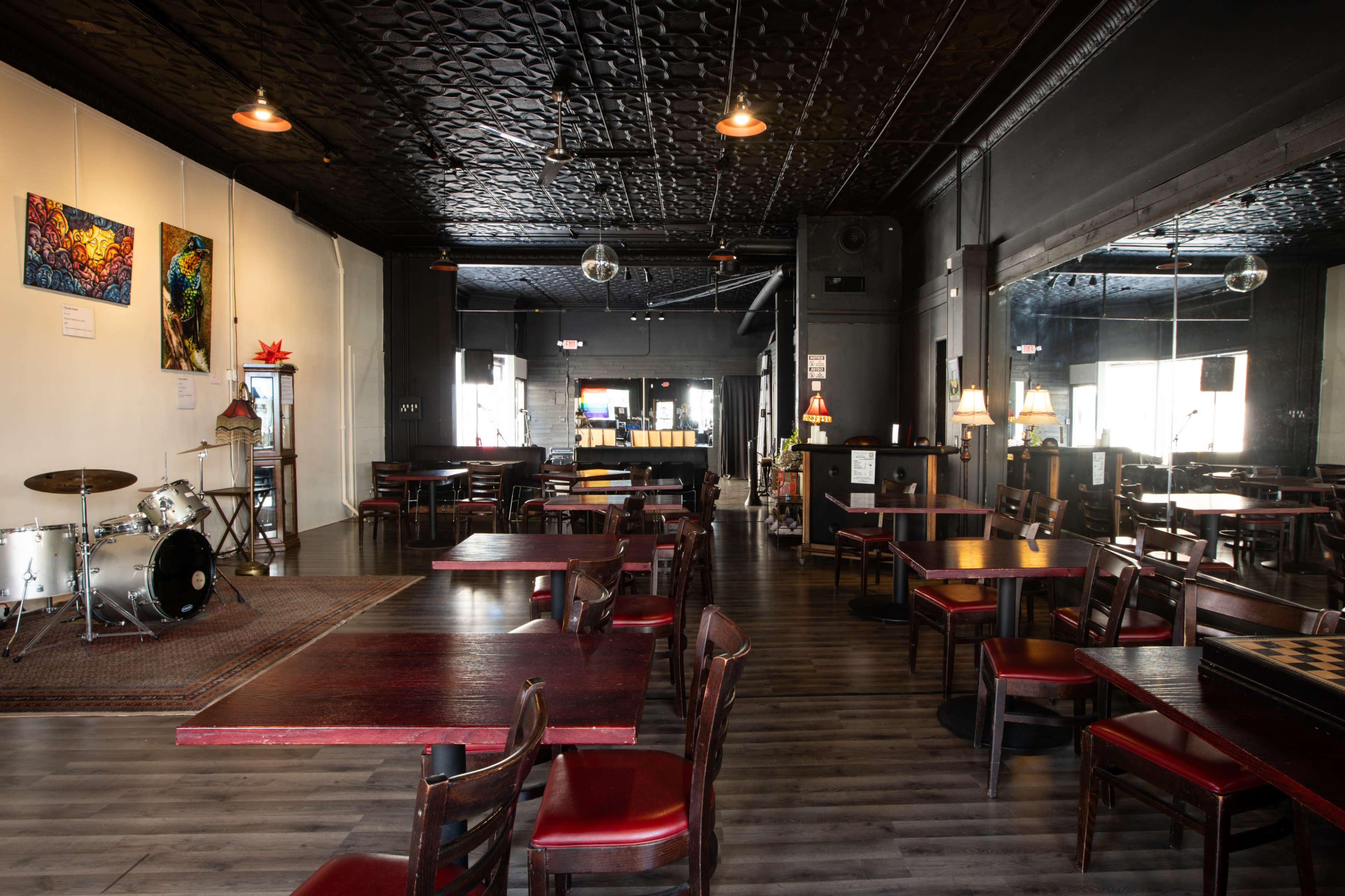The interior of a restaurant features wooden tables and chairs, a stage with drums in one corner, and colorful artwork on the walls.