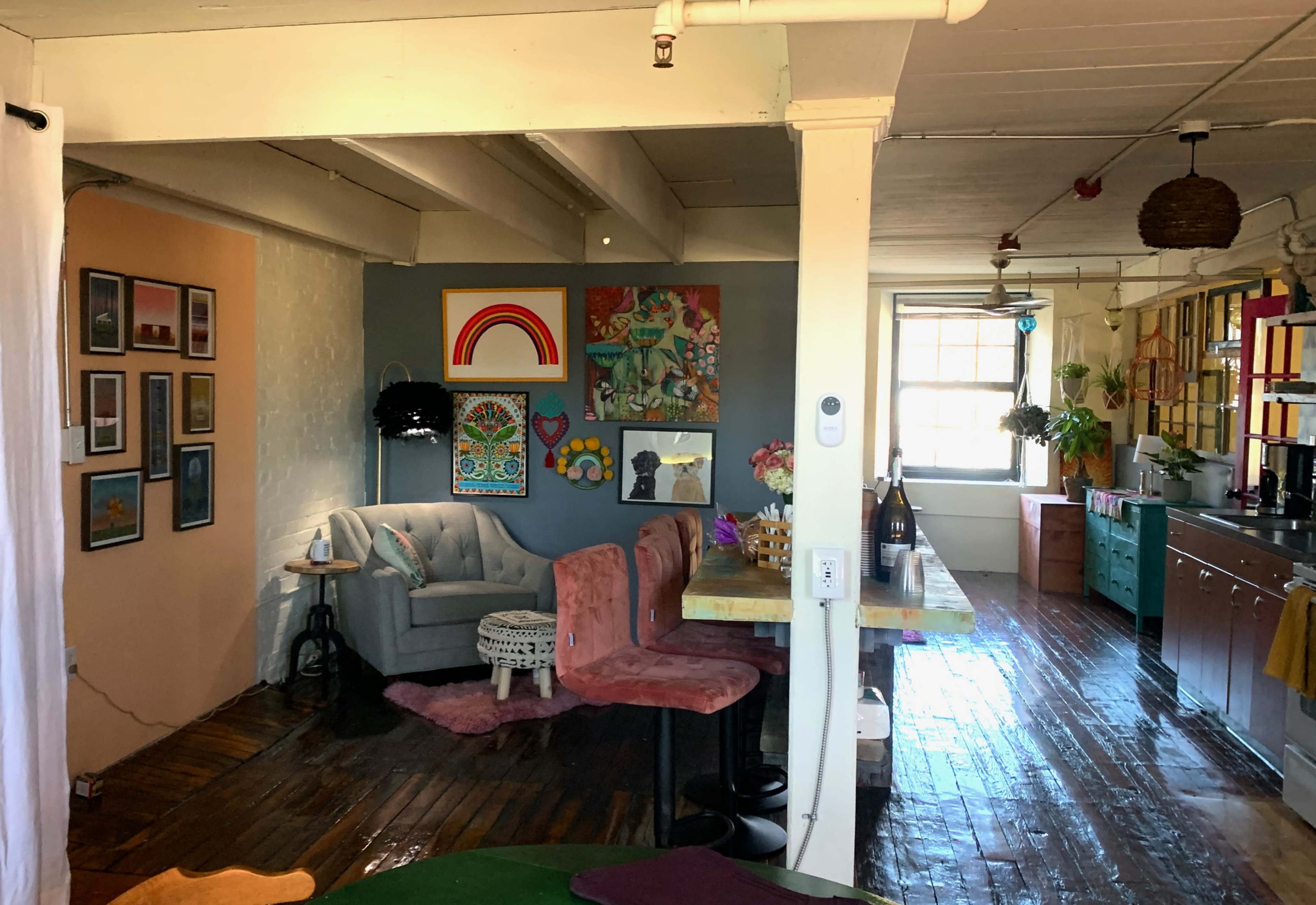 A cozy interior space with a living area featuring a gray armchair, colorful wall art, and a dining table within a bright room.