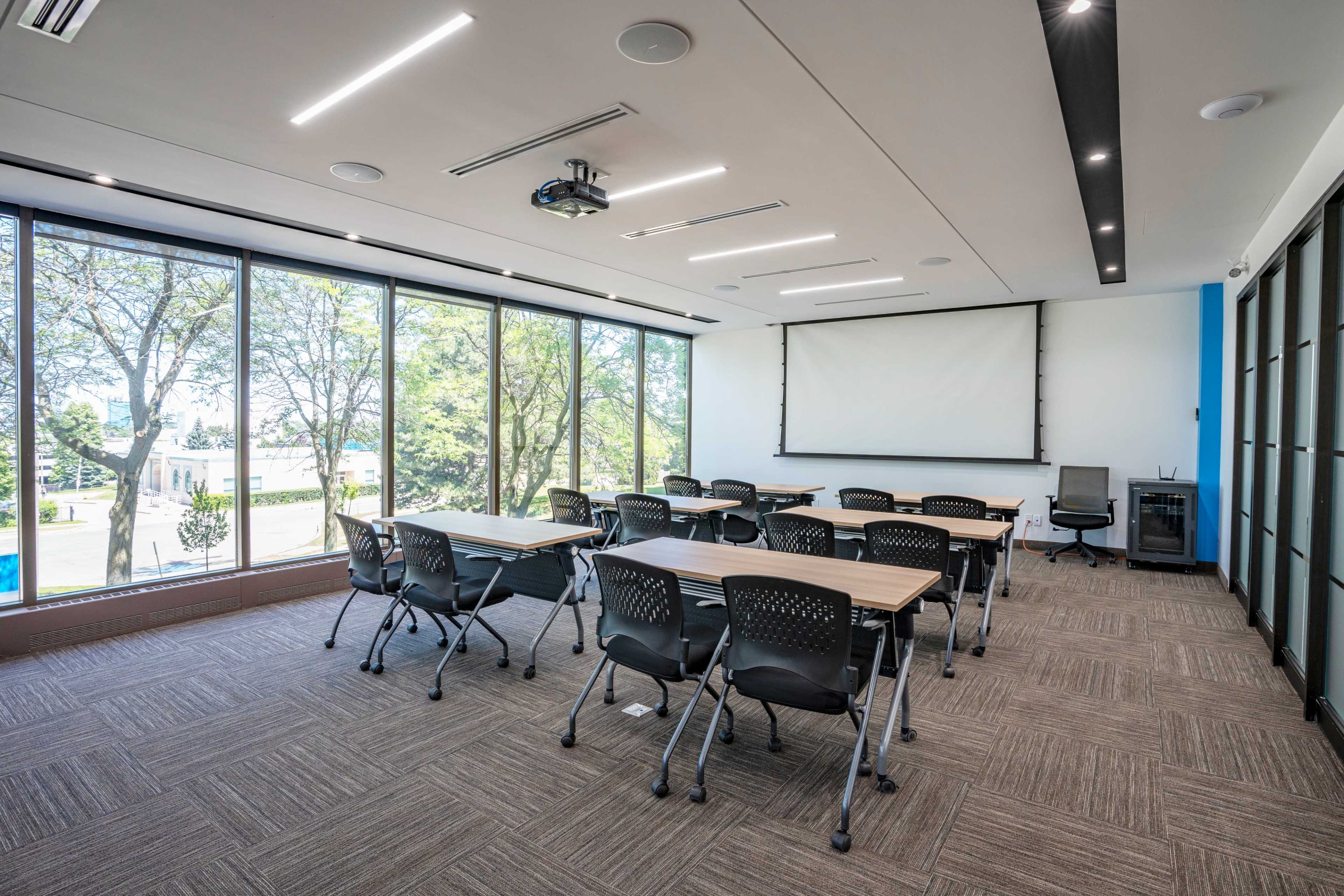 A modern conference room features a projector screen at the front, rows of tables and chairs, and large windows overlooking a green landscape.