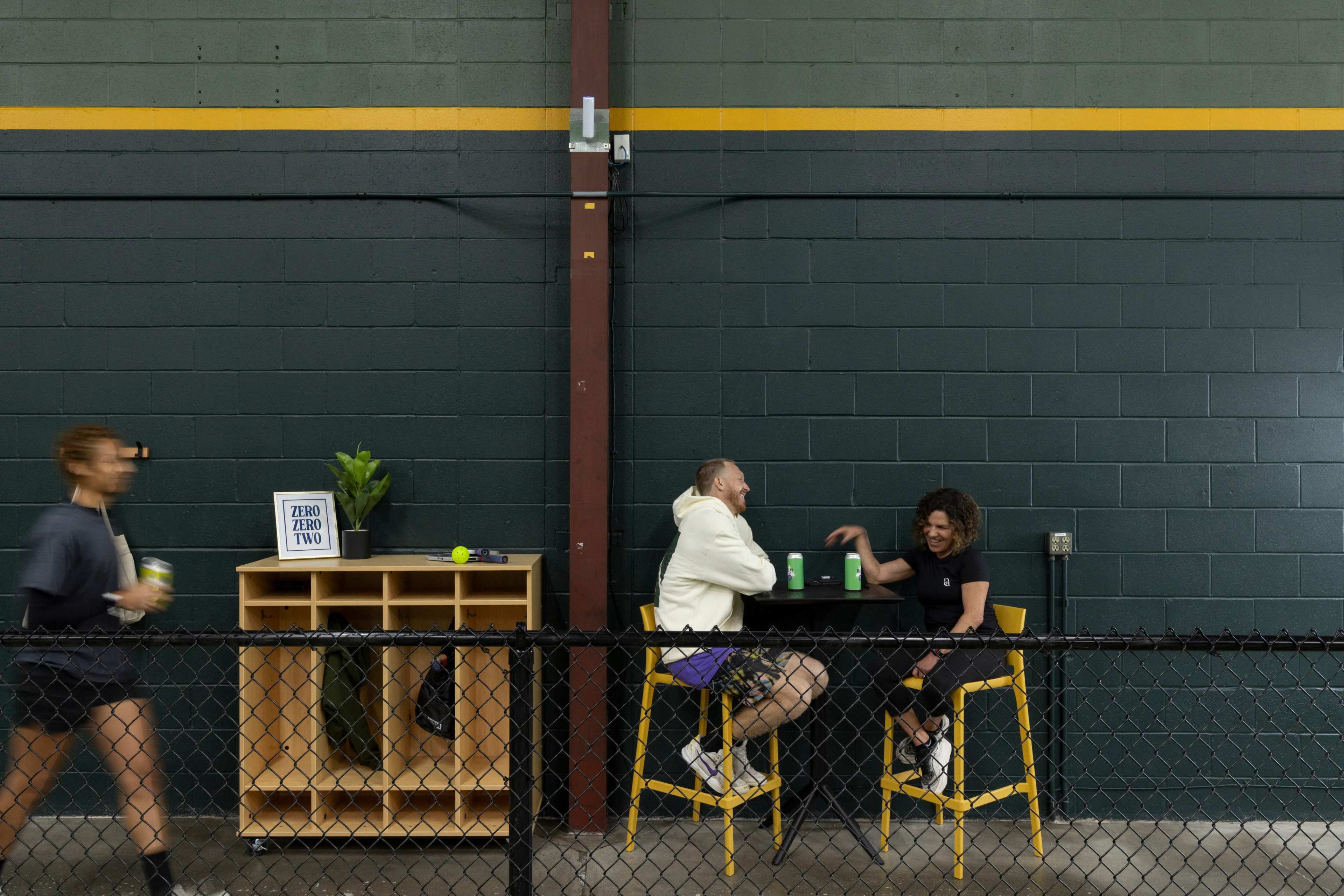 Two people sit at tall stools in a gym-like setting, engaged in conversation while a third person walks by holding a drink.
