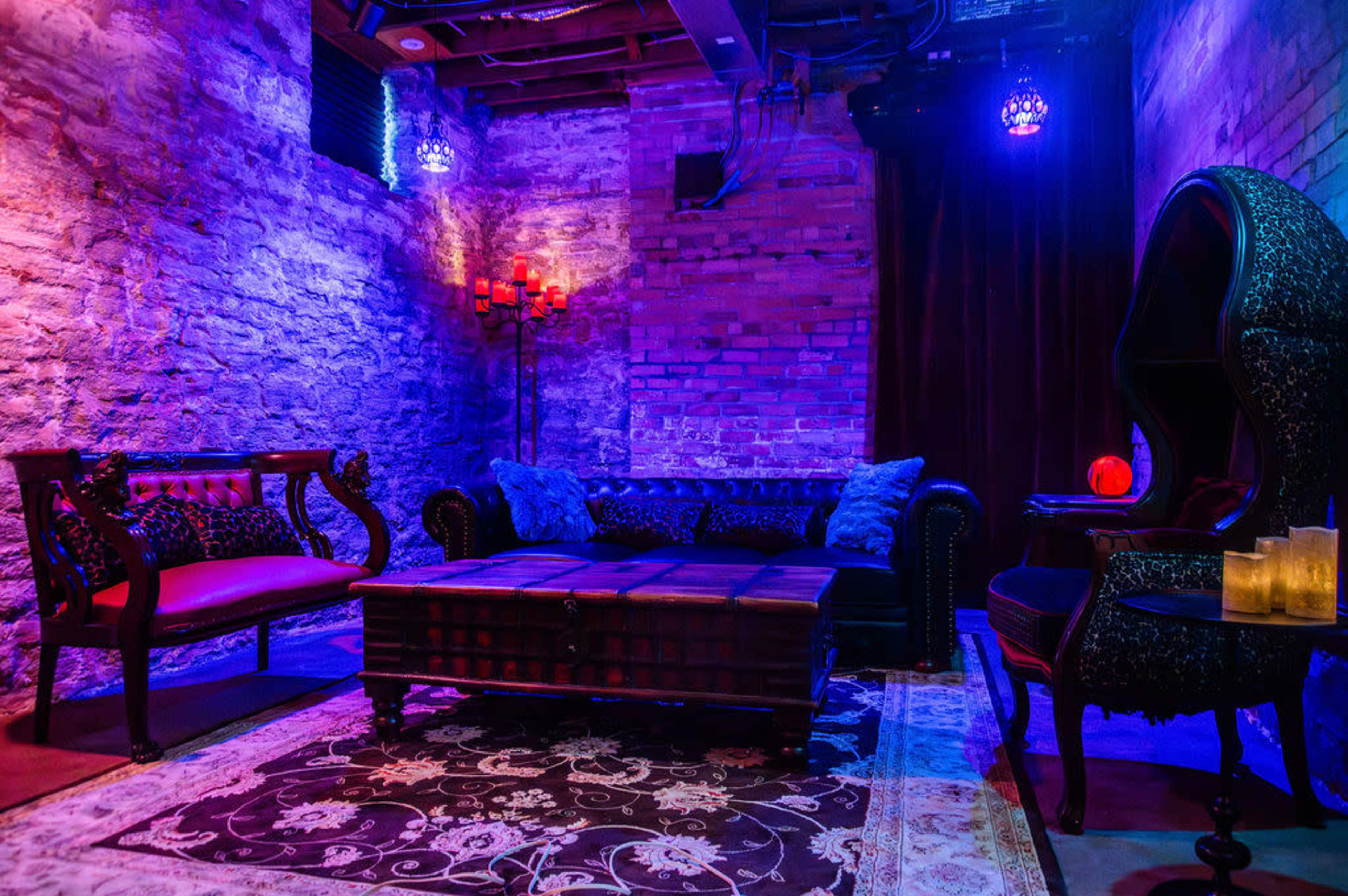 The image shows a dimly lit lounge area with a brick wall, dark furniture, and decorative elements, including a table, a plush rug, and candles.