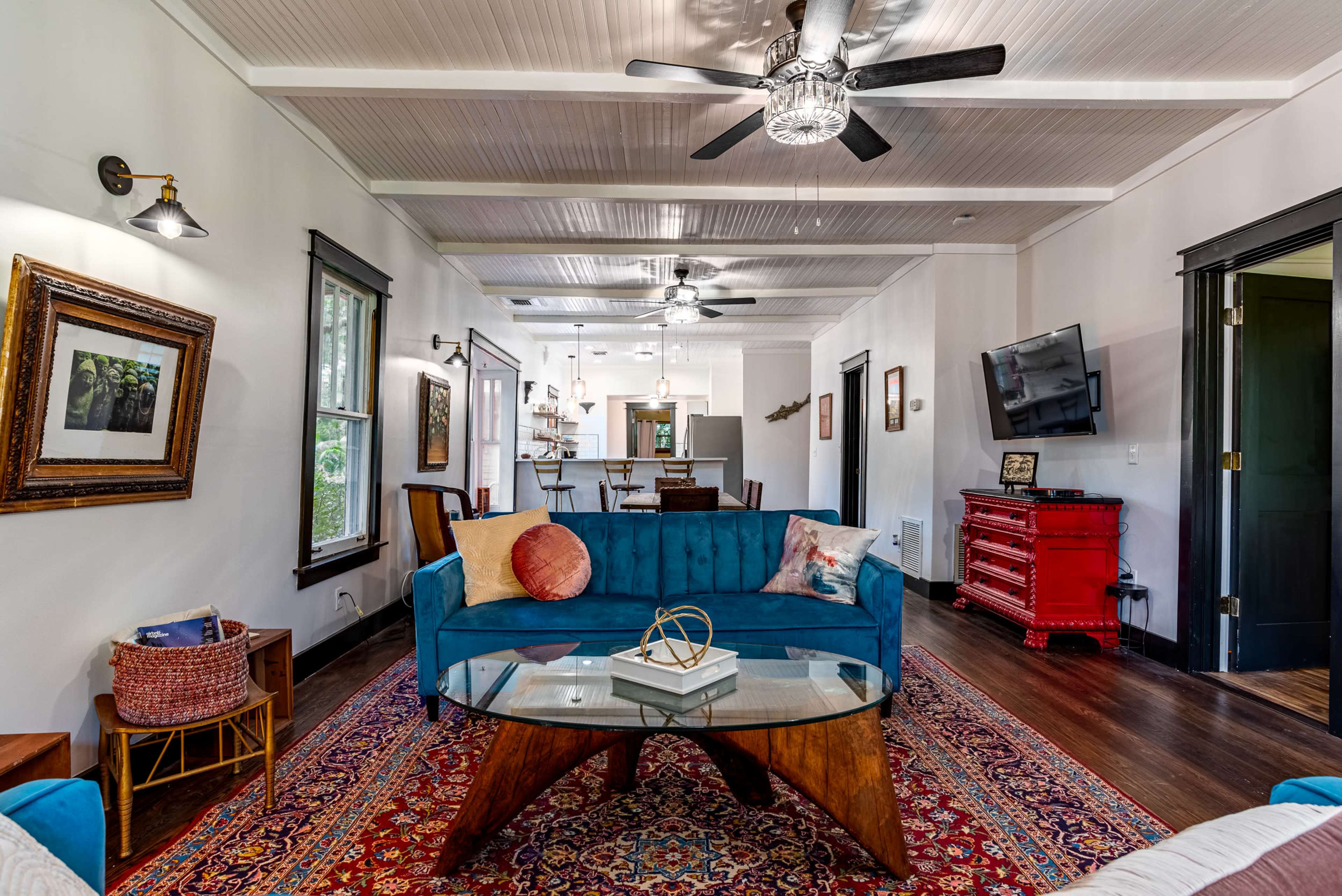 A living room featuring a blue velvet sofa, a glass-top coffee table, and a patterned rug, with an open kitchen visible in the background.