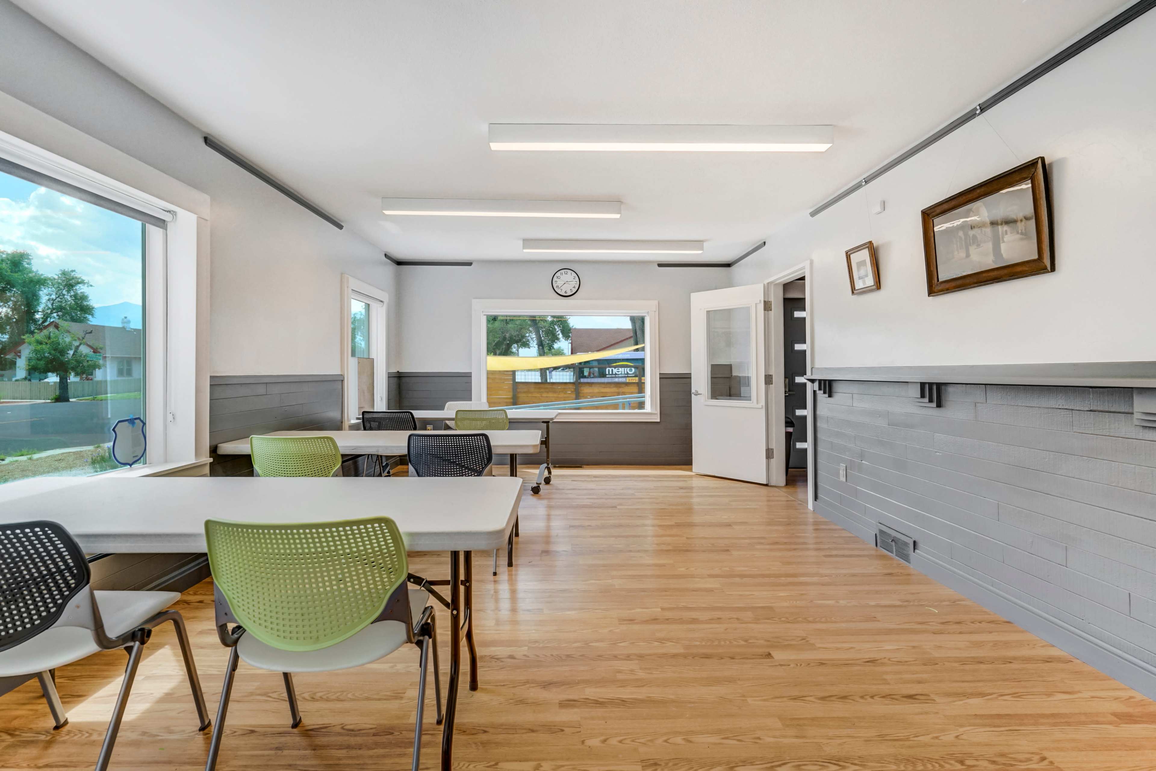 A bright classroom with several tables and chairs arranged neatly, large windows letting in light, and a clock on the wall.