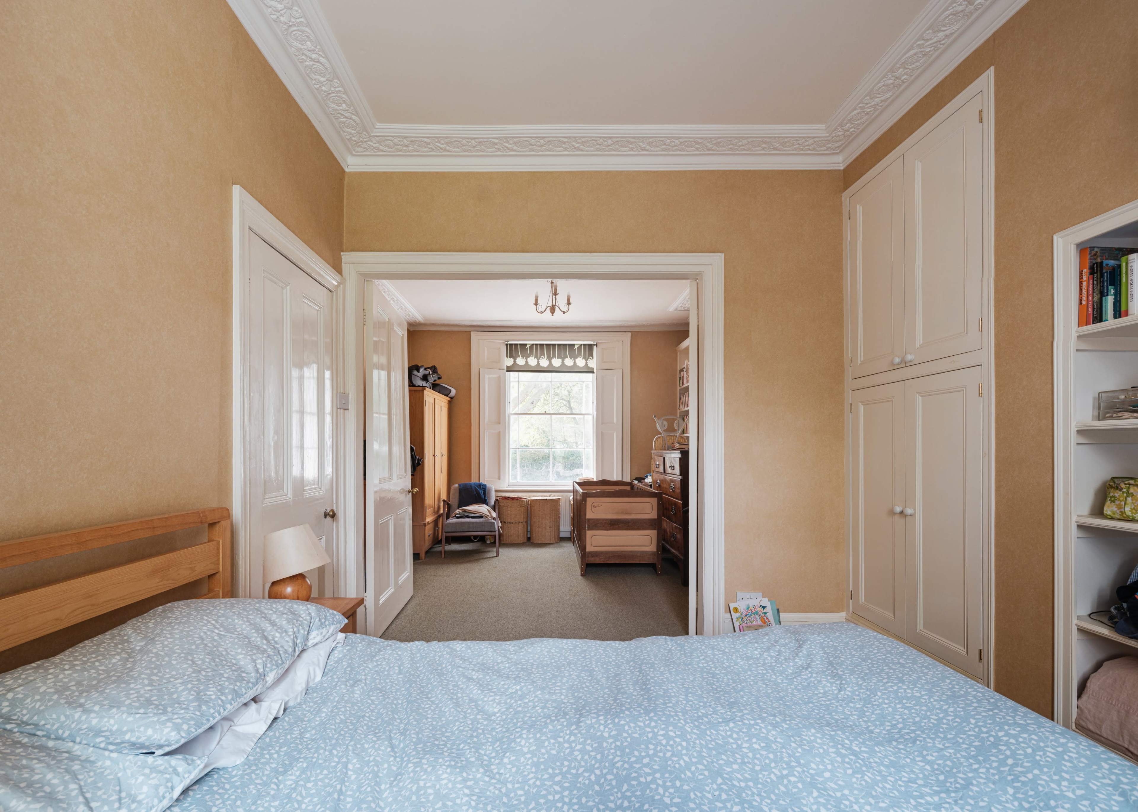A neatly arranged bedroom features a bed with a light blue cover, leading into a bright room with a window and various furniture pieces.