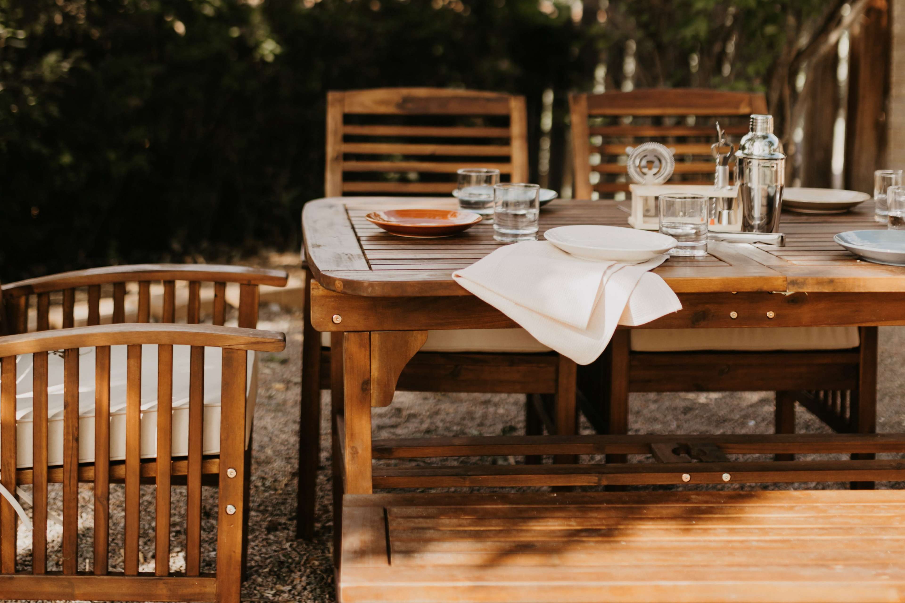 A wooden dining table with chairs is set outdoors with plates, glasses, and utensils arranged on it.