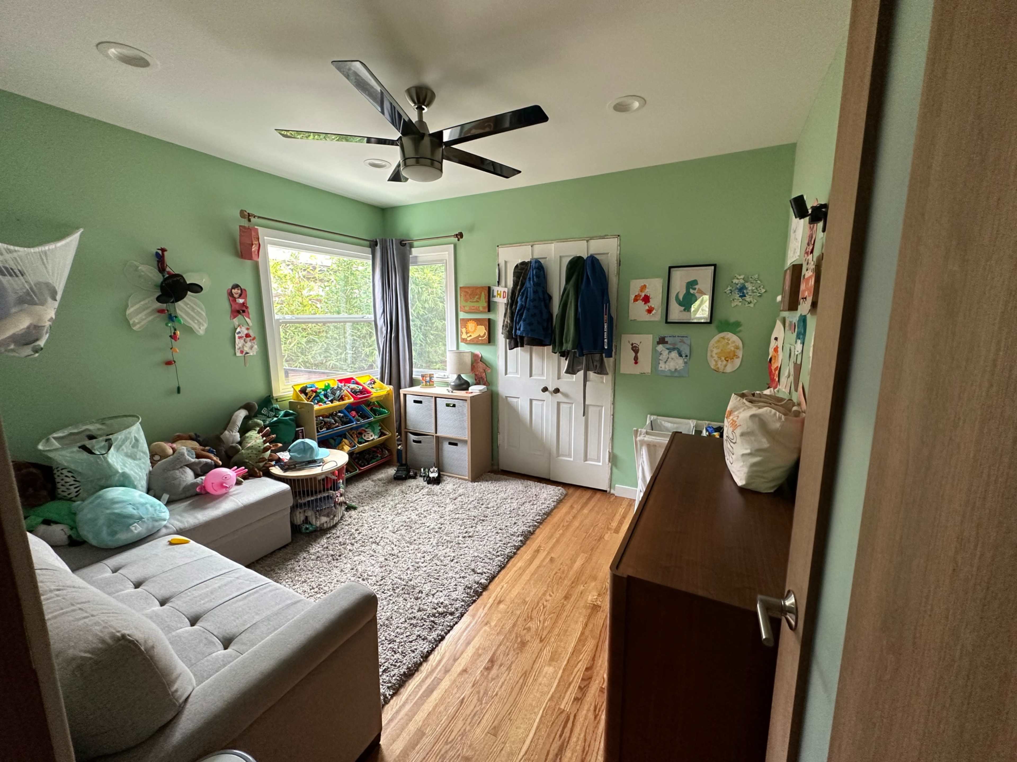A small, light green room with a sofa, a storage cabinet, and various toys scattered around, along with a window and a door.