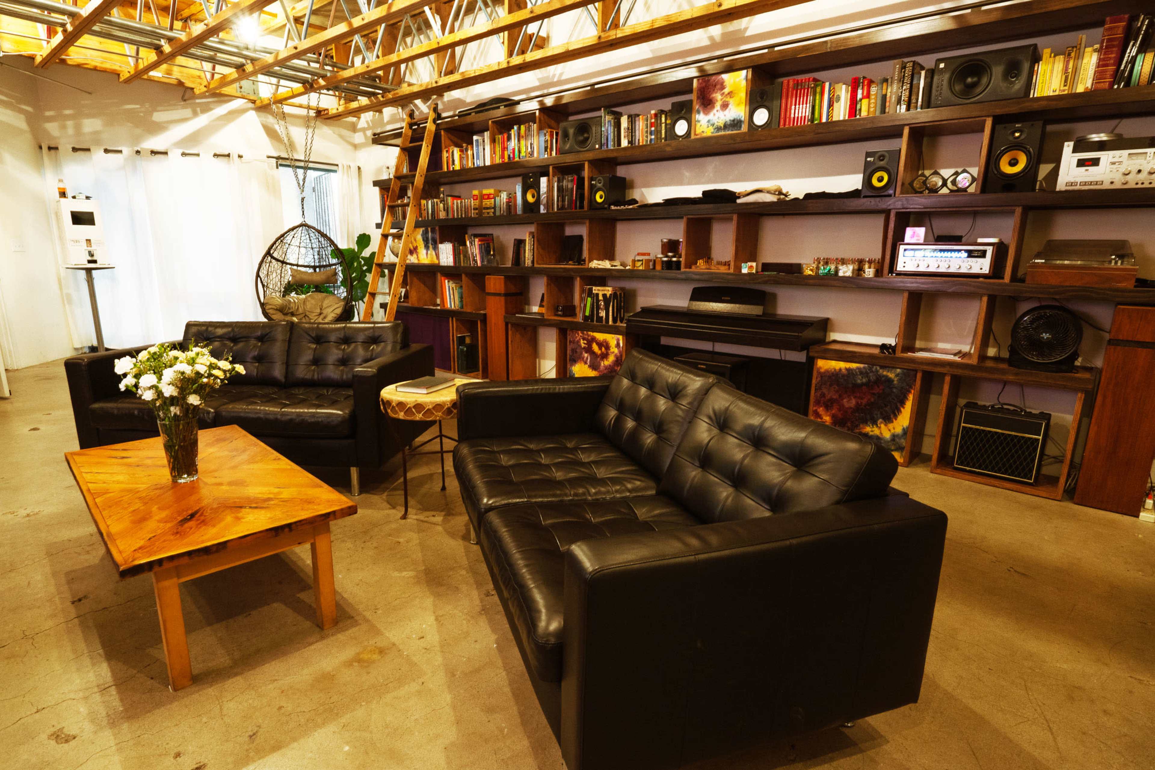 A cozy interior space with black leather sofas, a wooden coffee table, and a bookshelf filled with various items, illuminated by overhead lights.