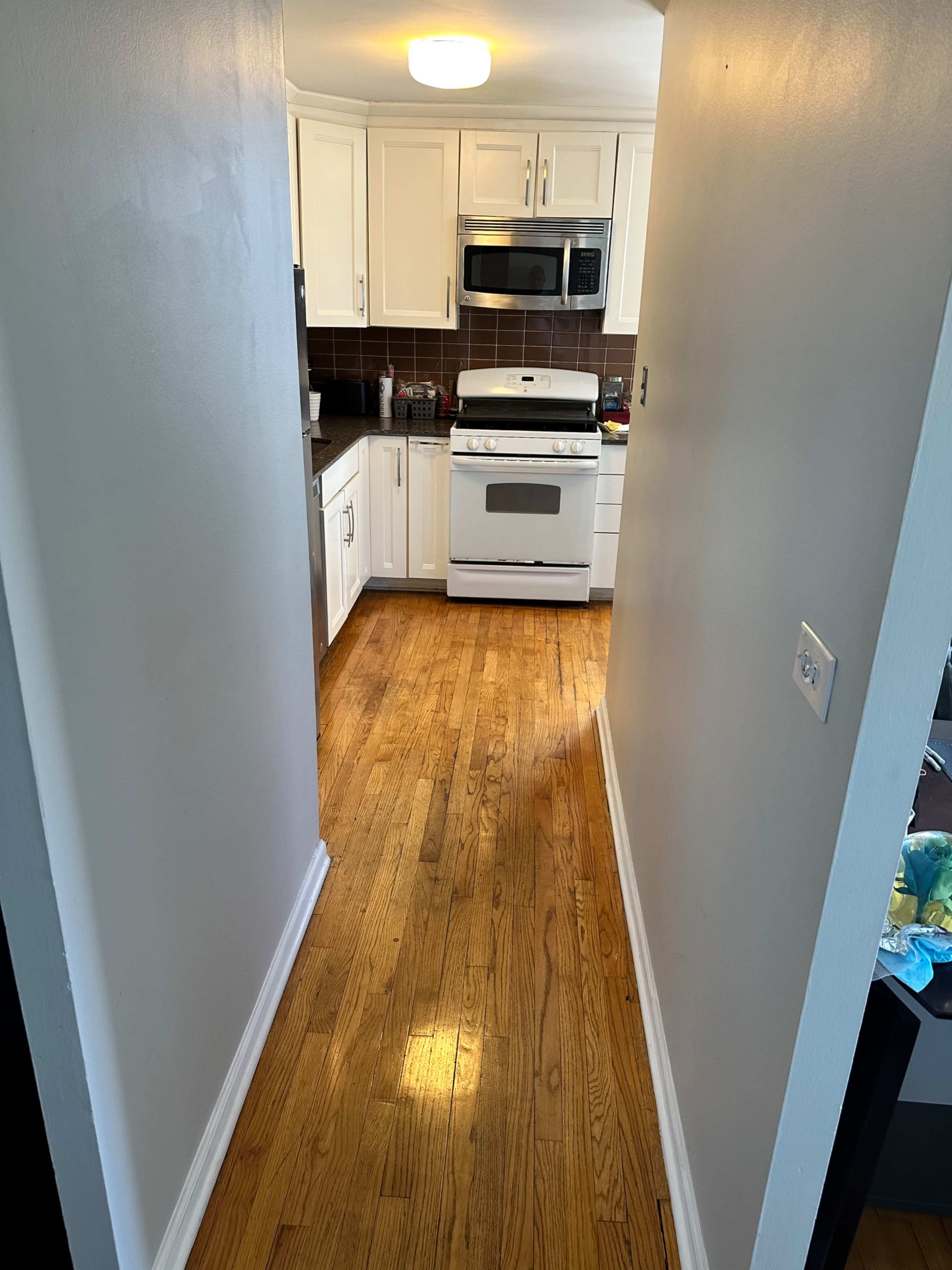 The image shows a narrow hallway leading into a kitchen with wooden flooring and white cabinetry.
