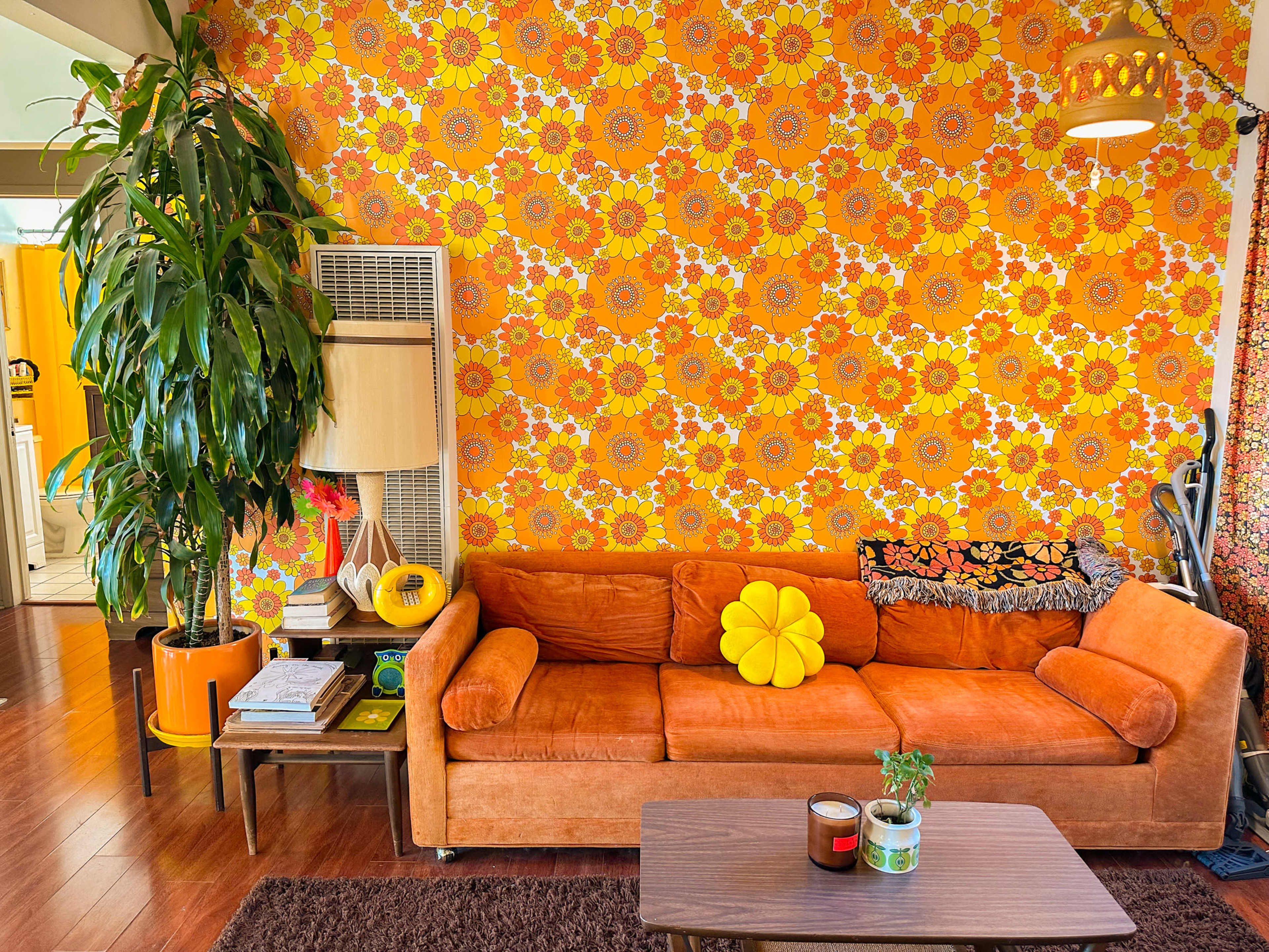 The image shows a living room with a large orange couch, a vintage lamp, a small coffee table, and a vibrant floral wallpaper in shades of orange and yellow.