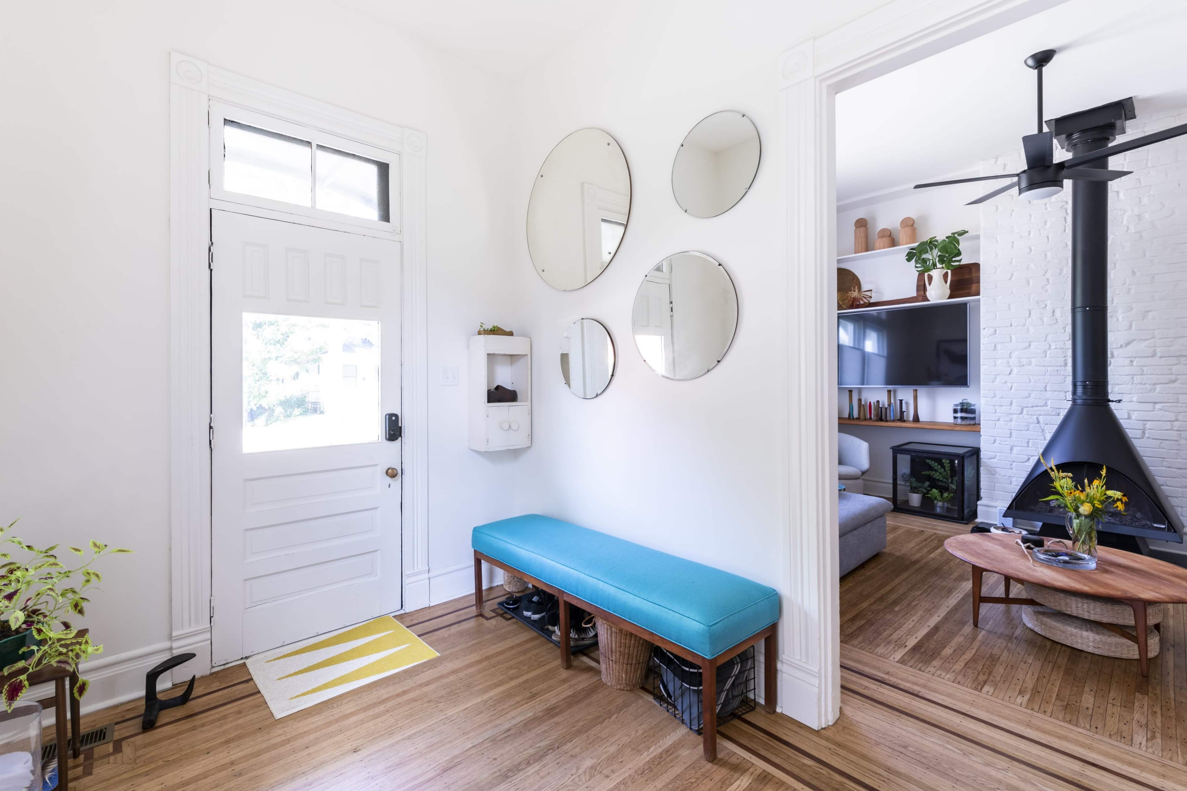 A modern entryway with a blue bench, circular mirrors on the wall, and a view into a living area featuring a black fireplace and light-colored furnishings.