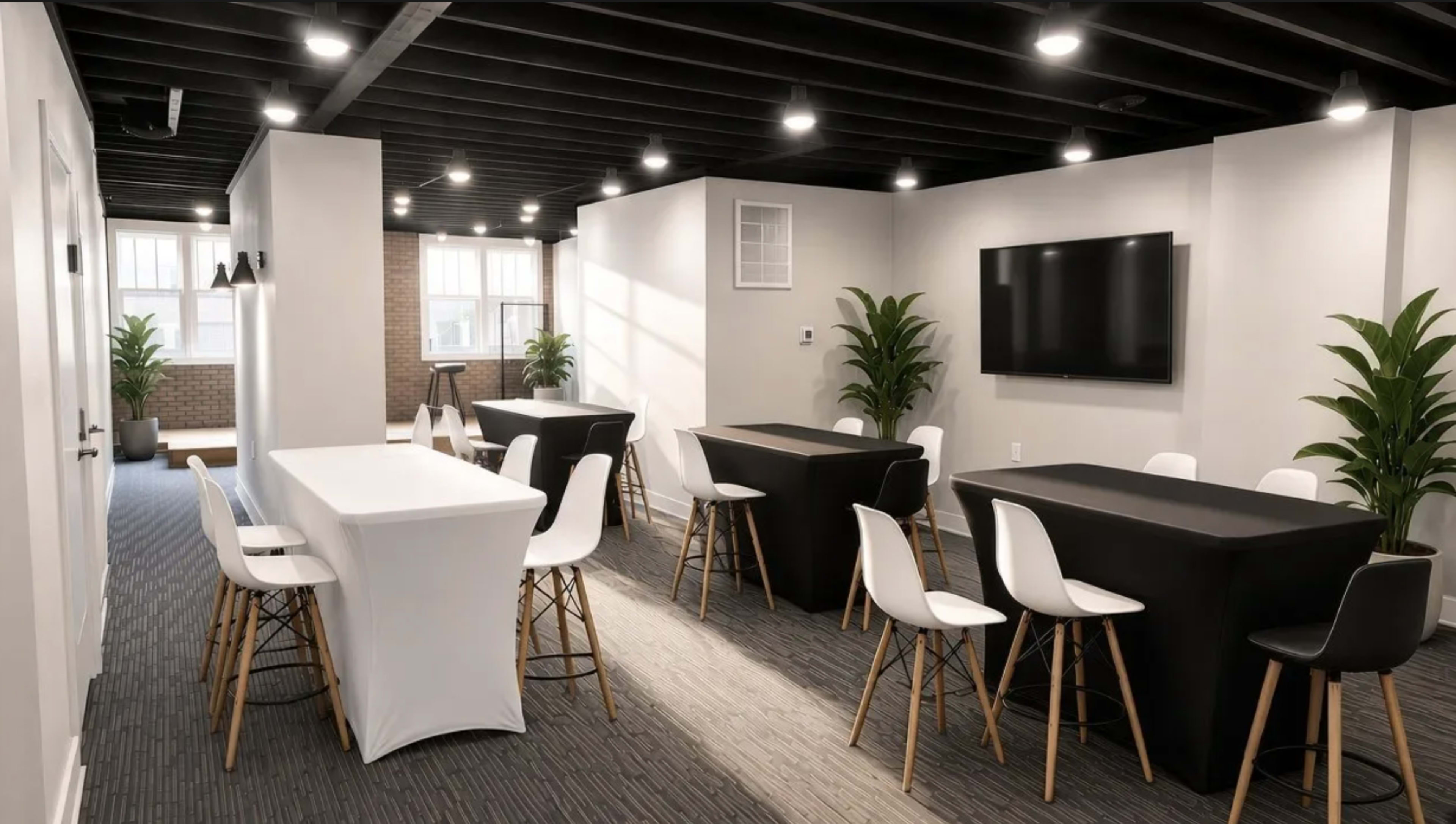 The image shows a bright, modern room with black and white tables and chairs, illuminated by overhead lights, and featuring potted plants in each corner.