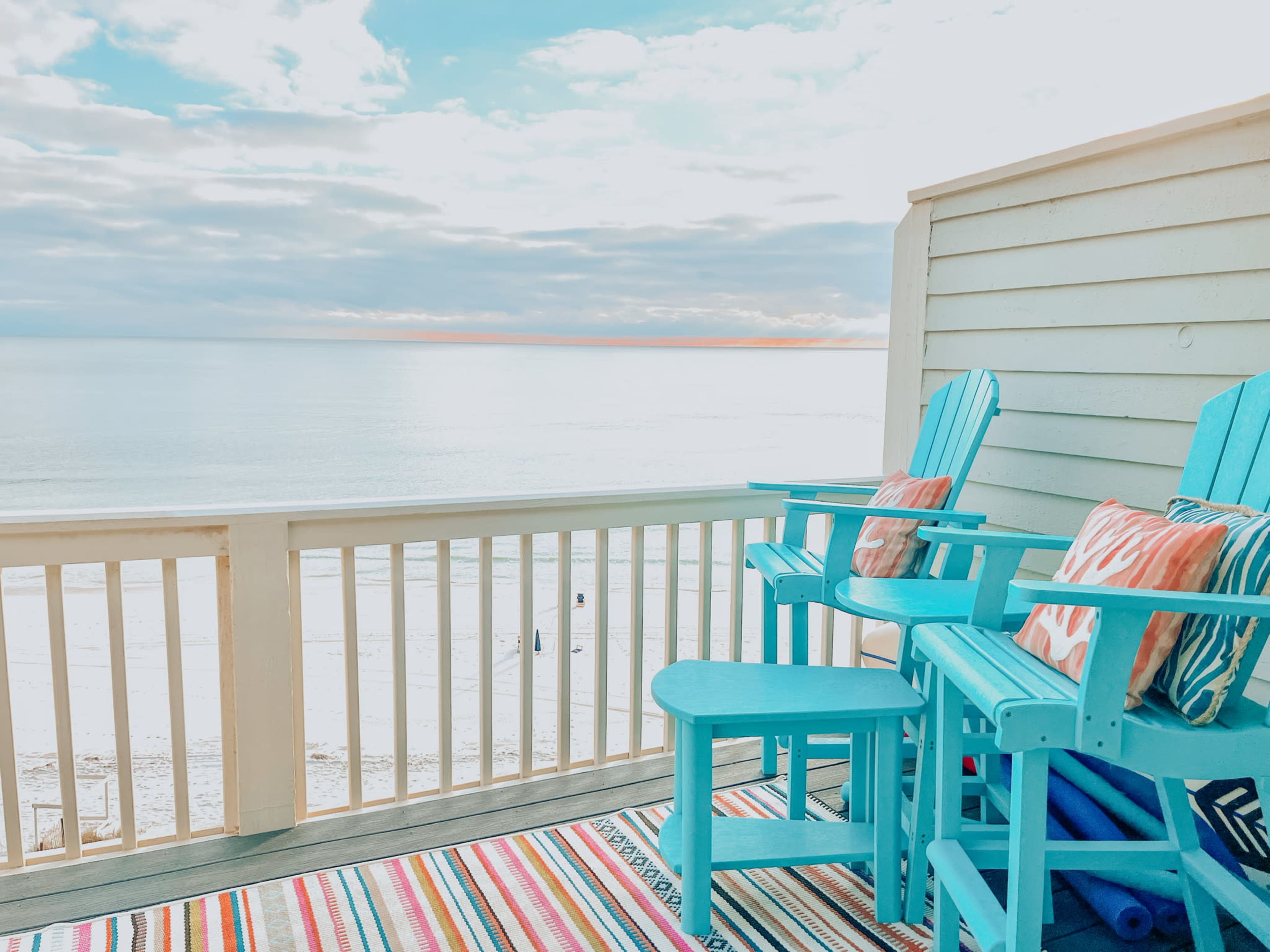 A balcony features two blue chairs and a small table overlooking a serene beach and calm ocean waters.