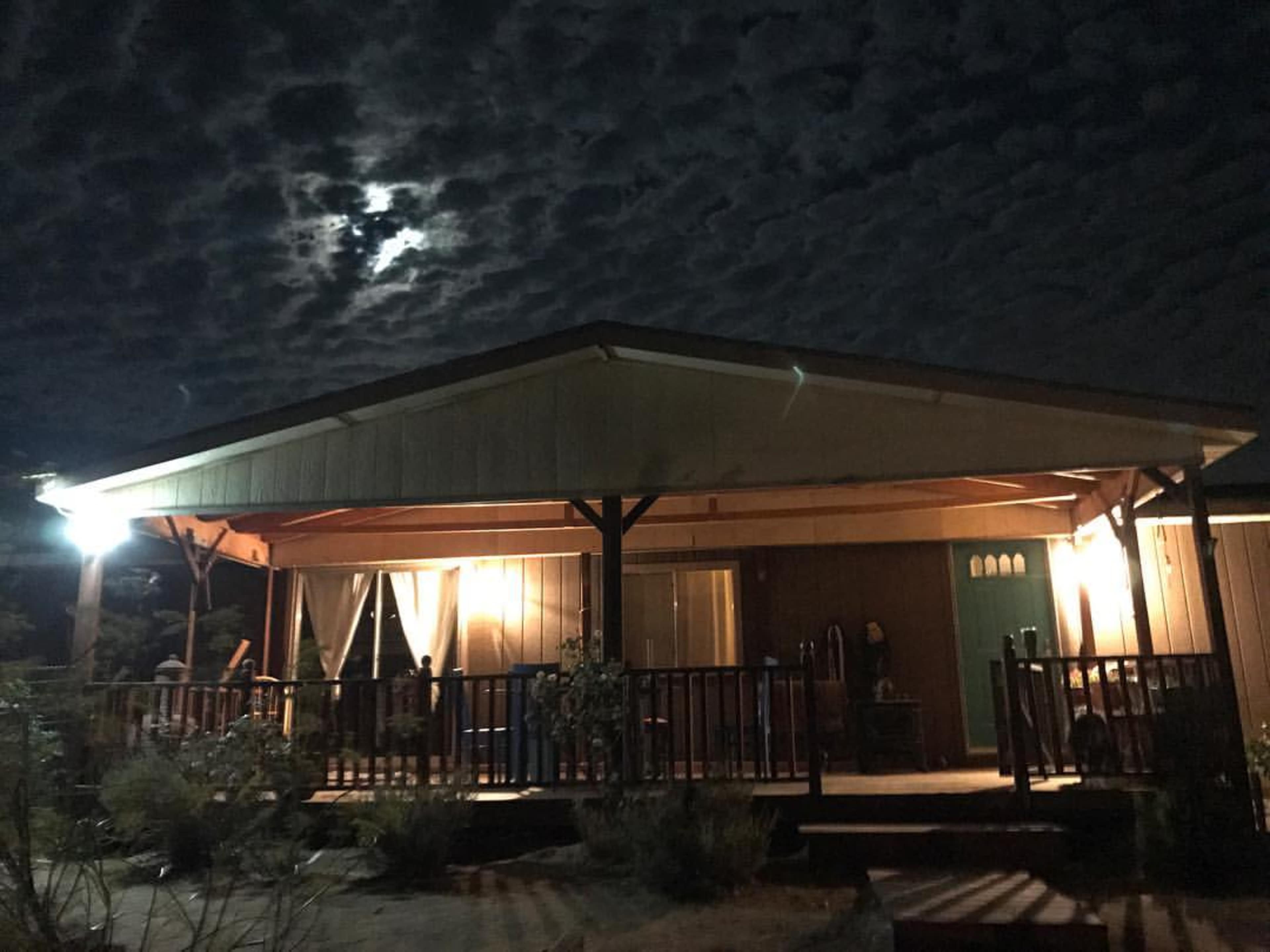 A wooden cabin with a porch is illuminated by artificial lights under a cloudy night sky.