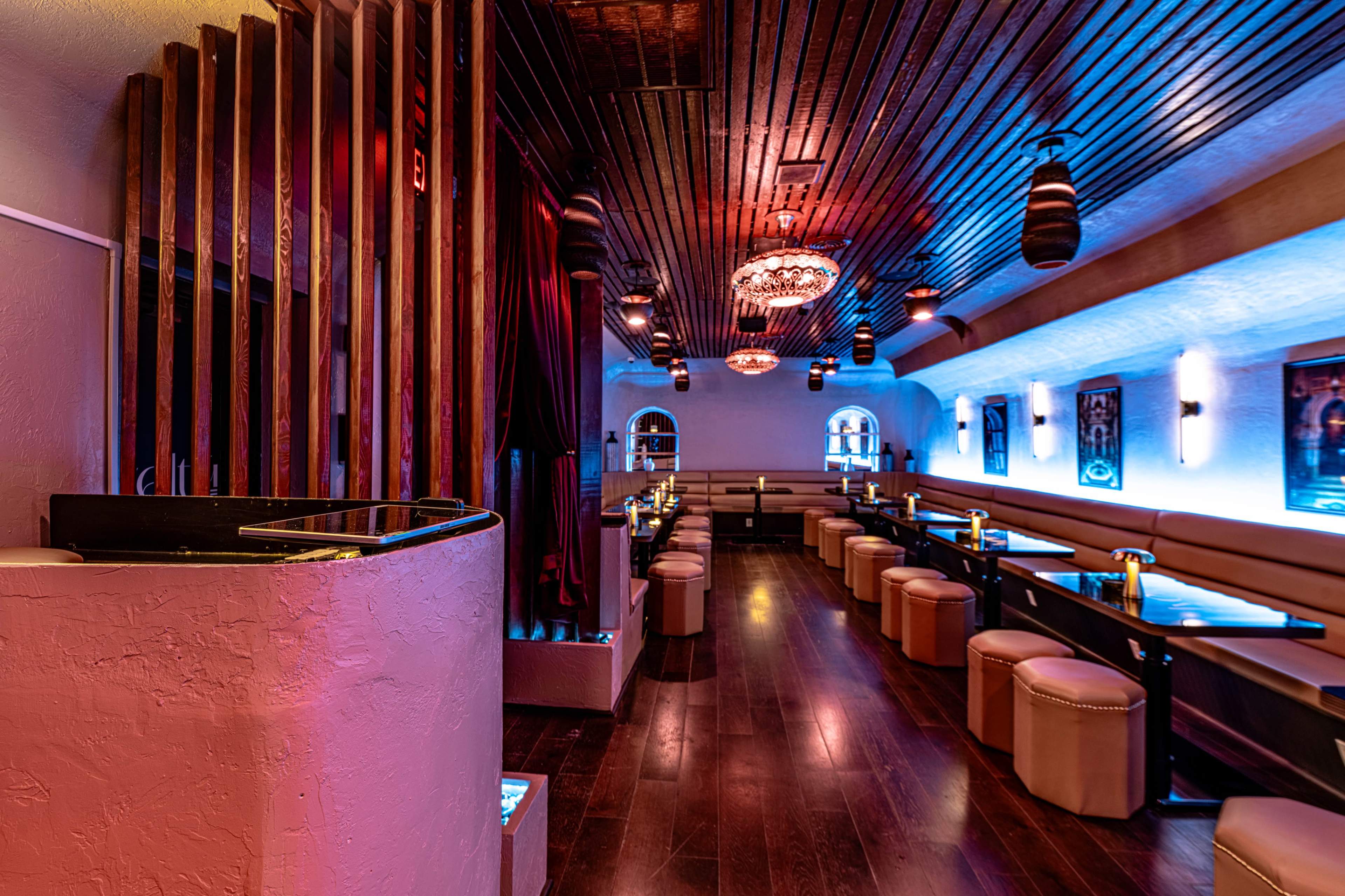 The image shows a modern restaurant interior featuring wooden ceiling beams, plush seating along the walls, and dim, colorful lighting.