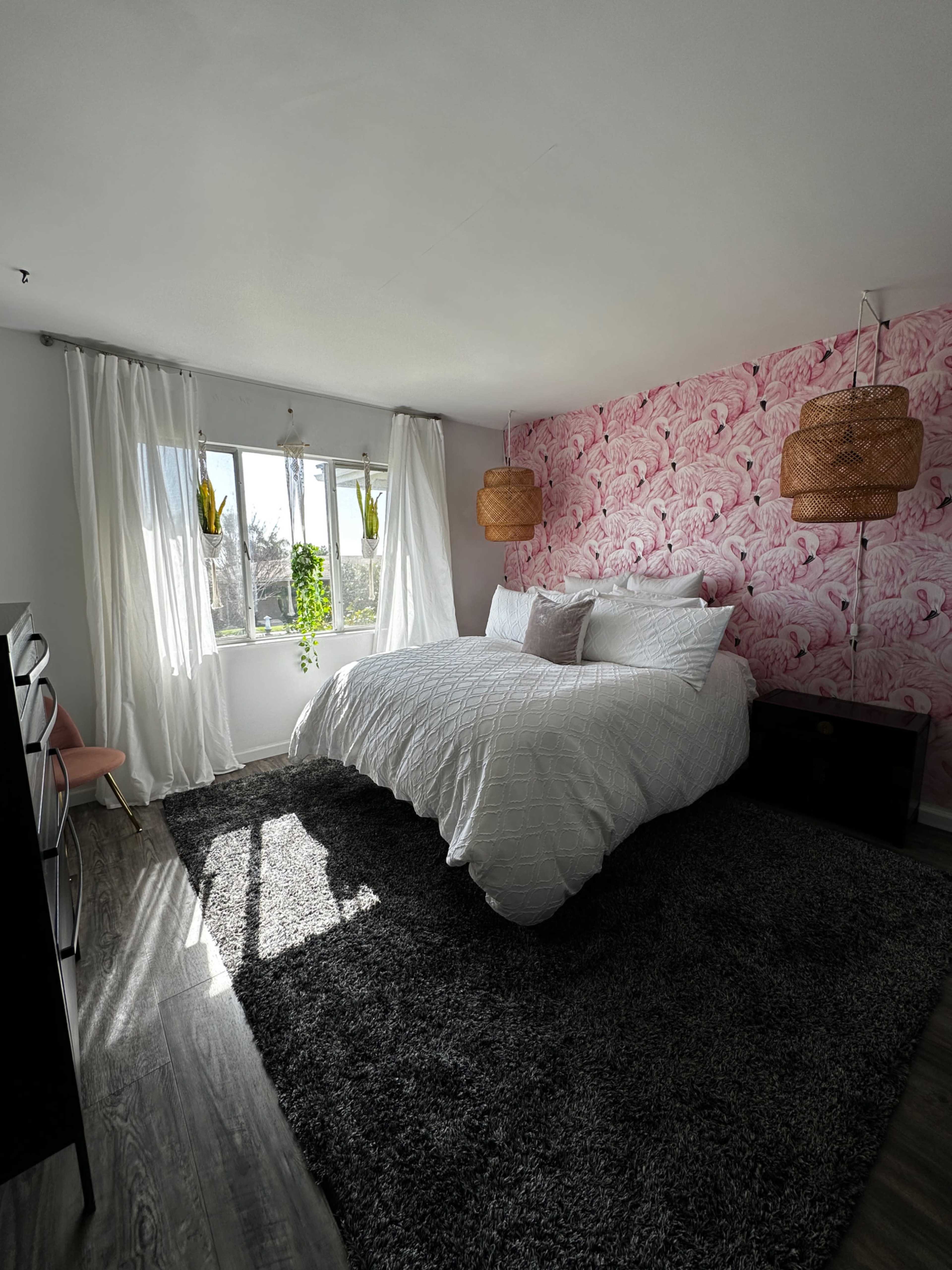 The bedroom features a bed with white bedding, a pink floral accent wall, large windows with sheer curtains, and woven pendant lights.