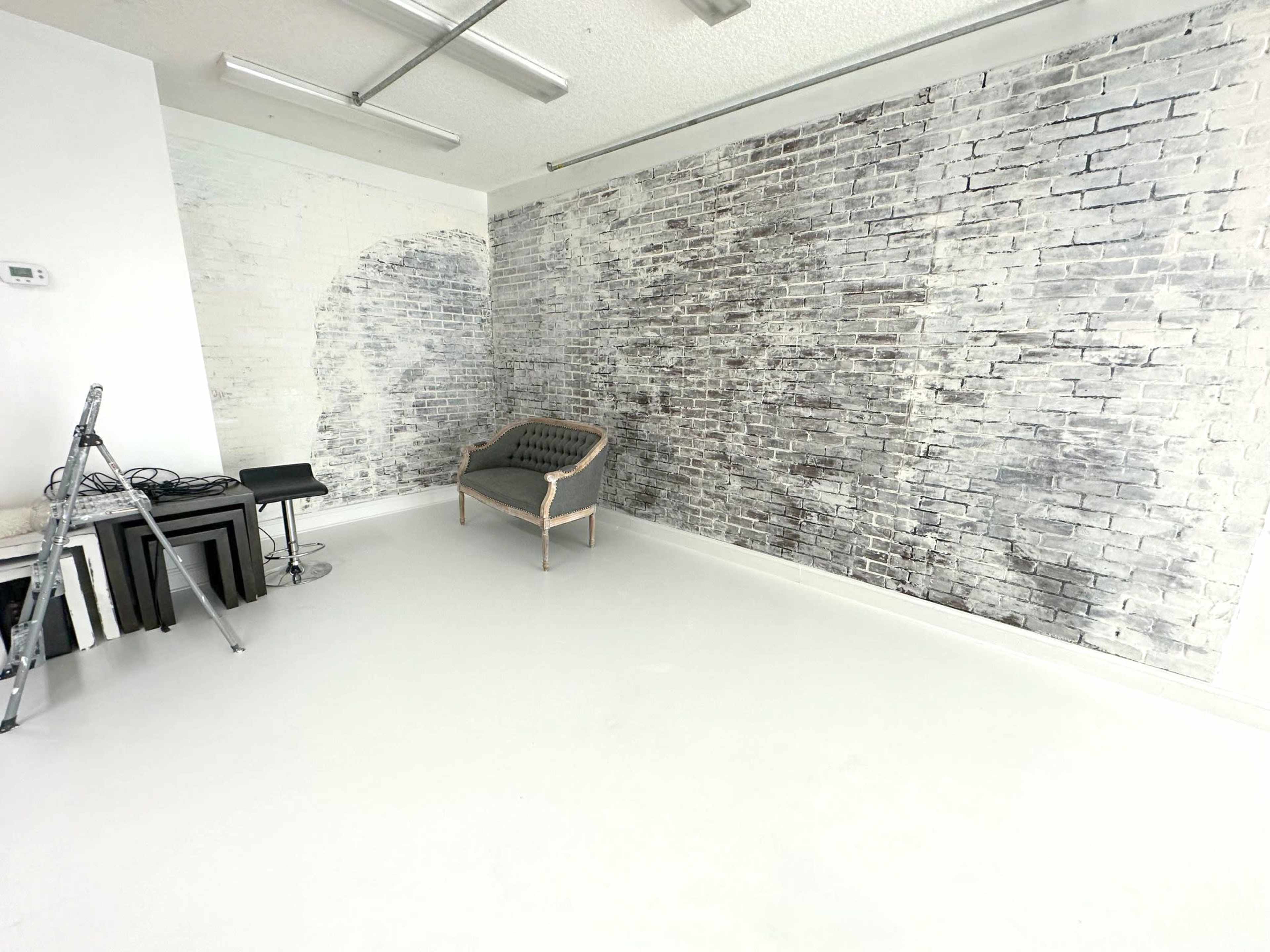 A room with a white floor, a sofa against a textured brick wall, and a few pieces of furniture including a ladder and side tables.