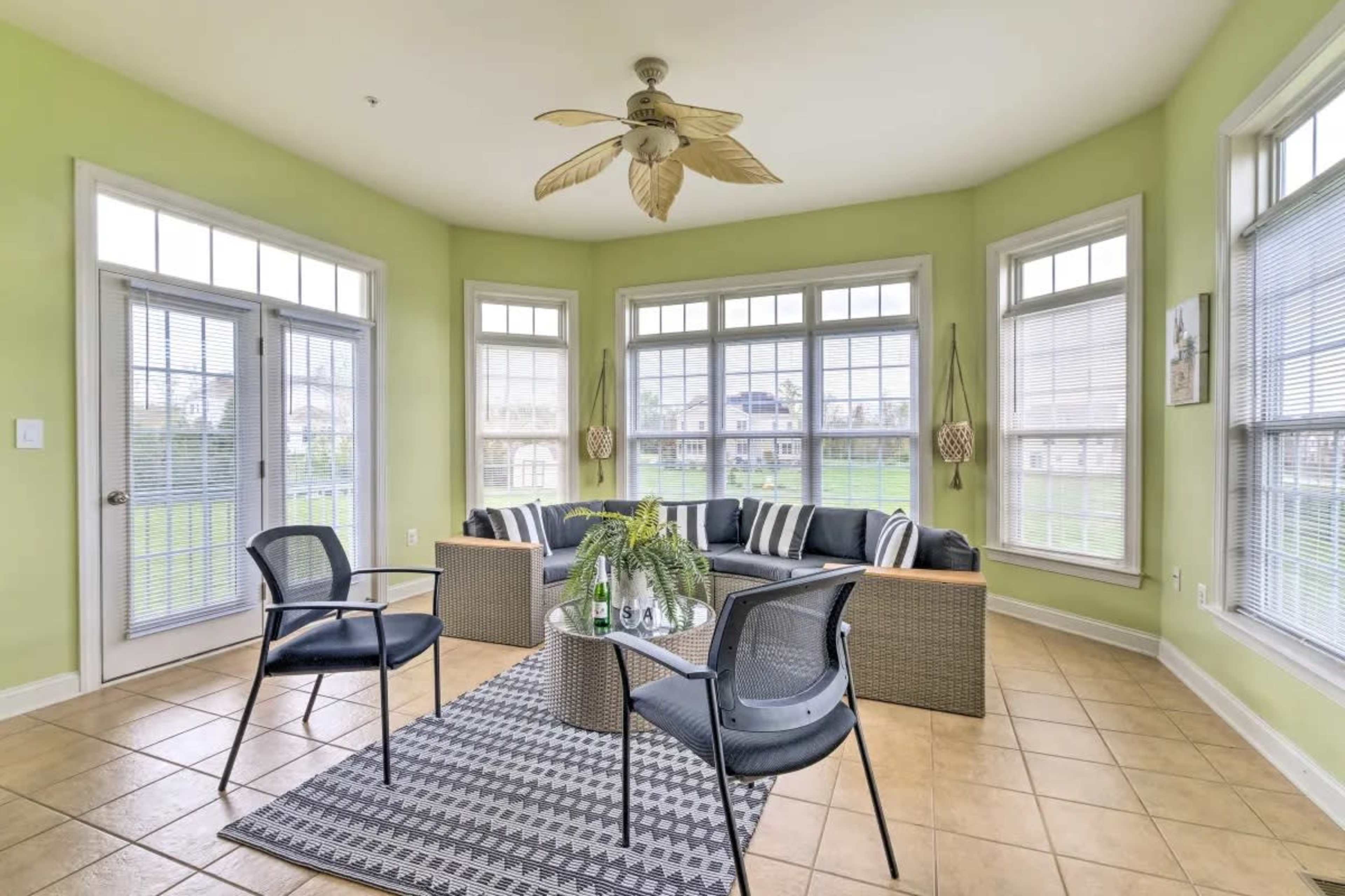 A sunlit sunroom features a rattan seating area with black cushions, surrounded by large windows and a patterned rug on tiled flooring.