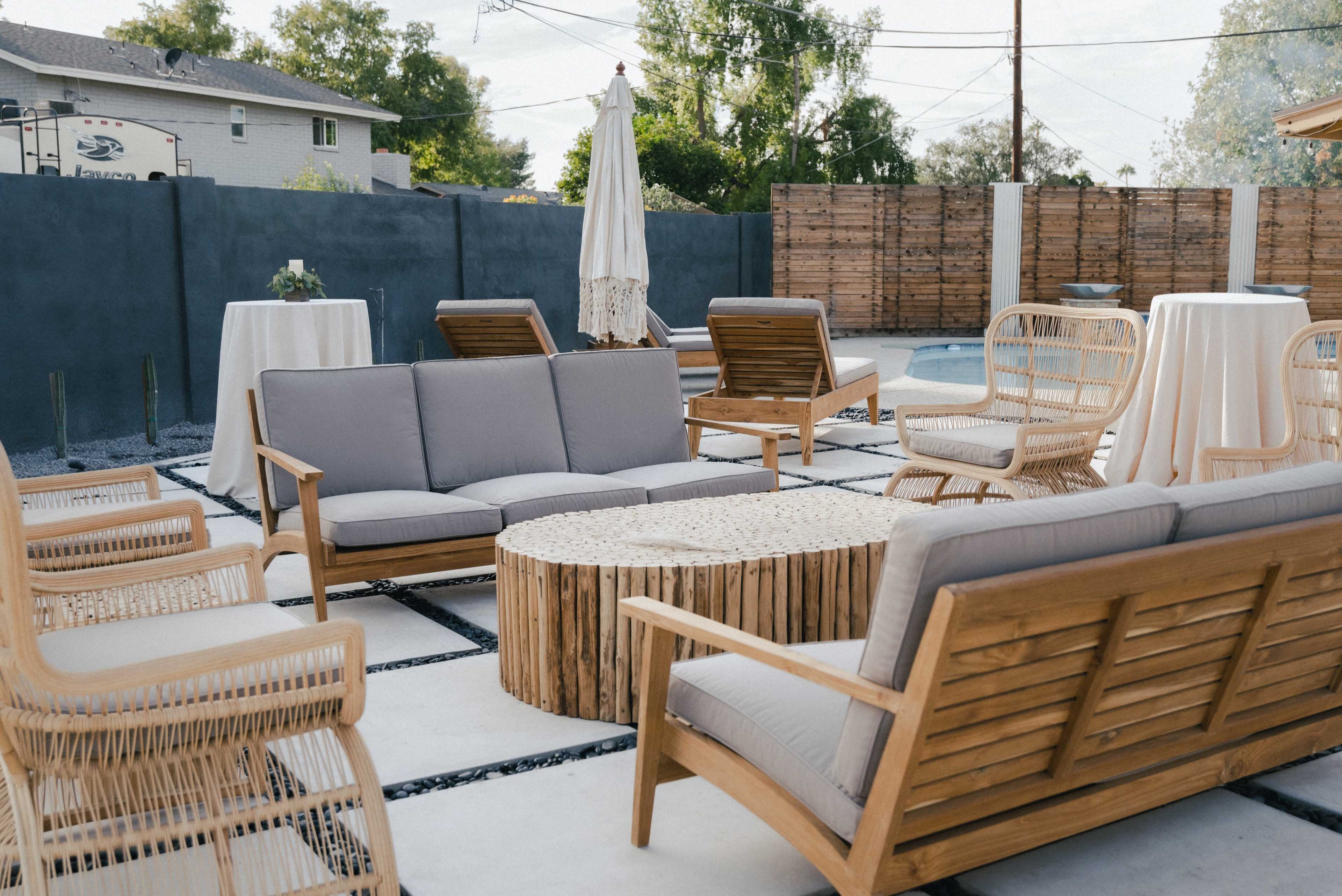 A patio area with wooden furniture, including a circular coffee table, surrounded by several chairs and two tall tables, in a setting with a pool and a wooden fence.