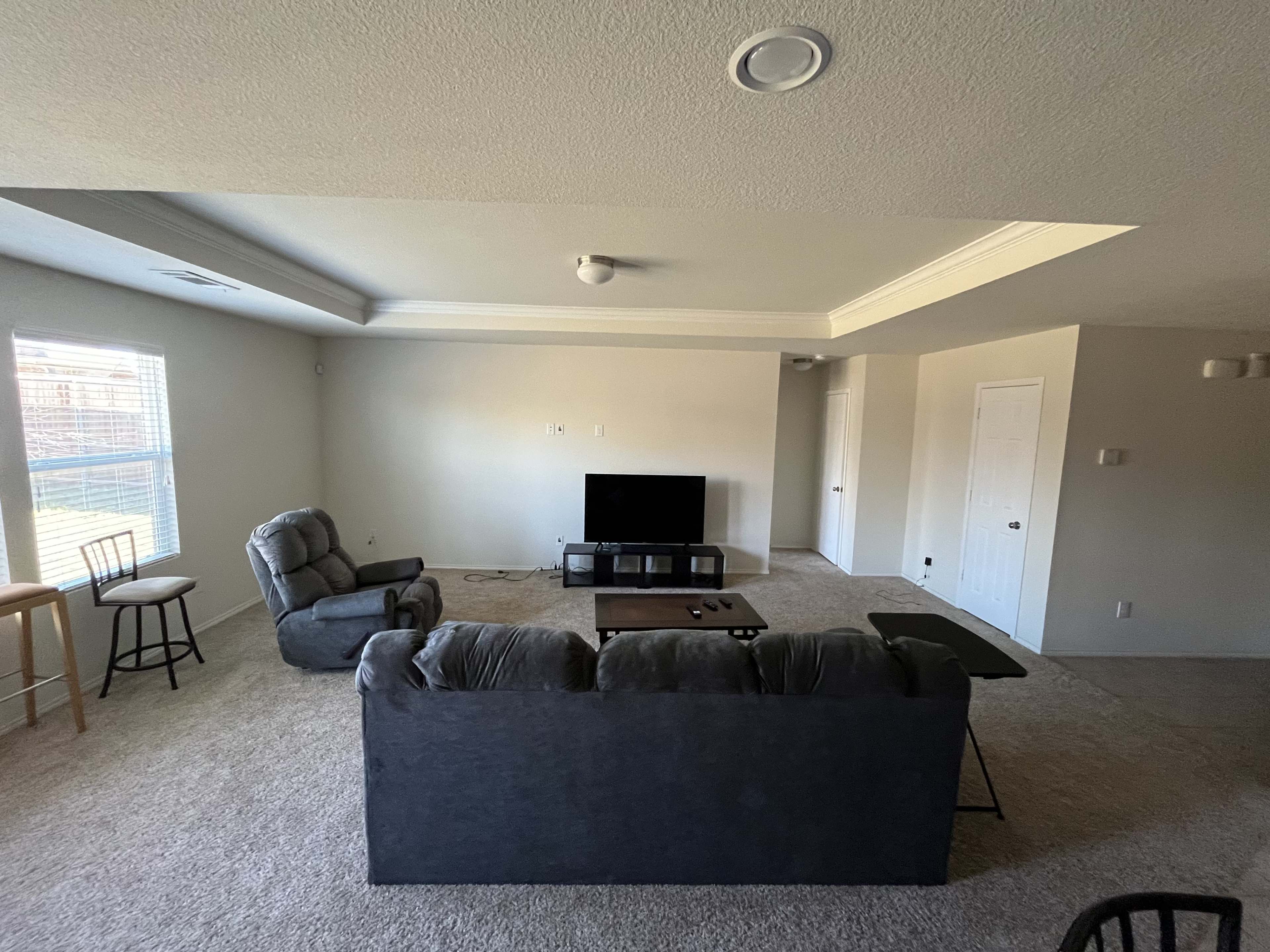 The image shows a spacious living room with a gray couch, two chairs, a television stand, and large windows allowing natural light to enter.