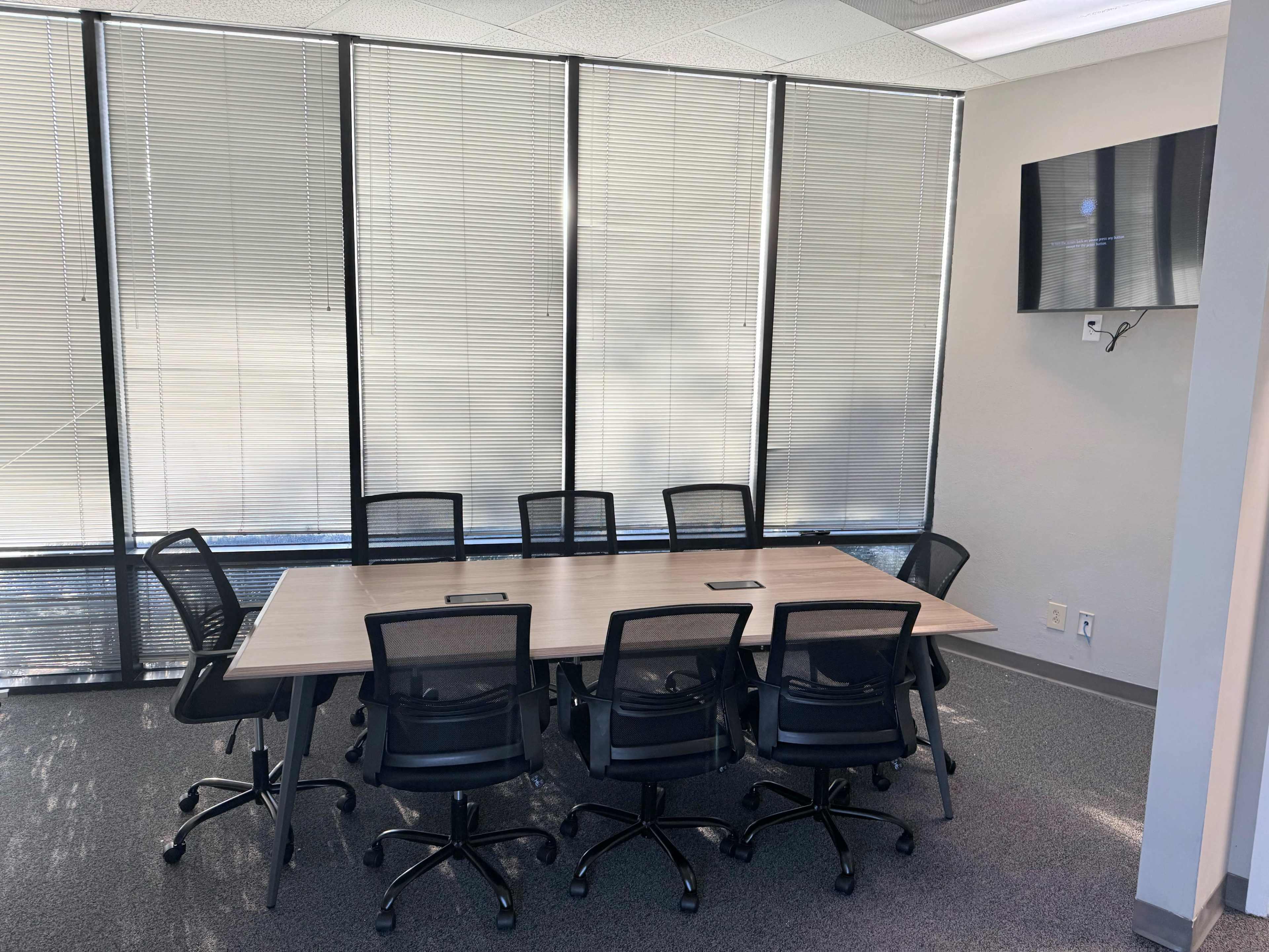 A modern conference room features a long table surrounded by eight black chairs, with large windows covered by blinds and a wall-mounted television.