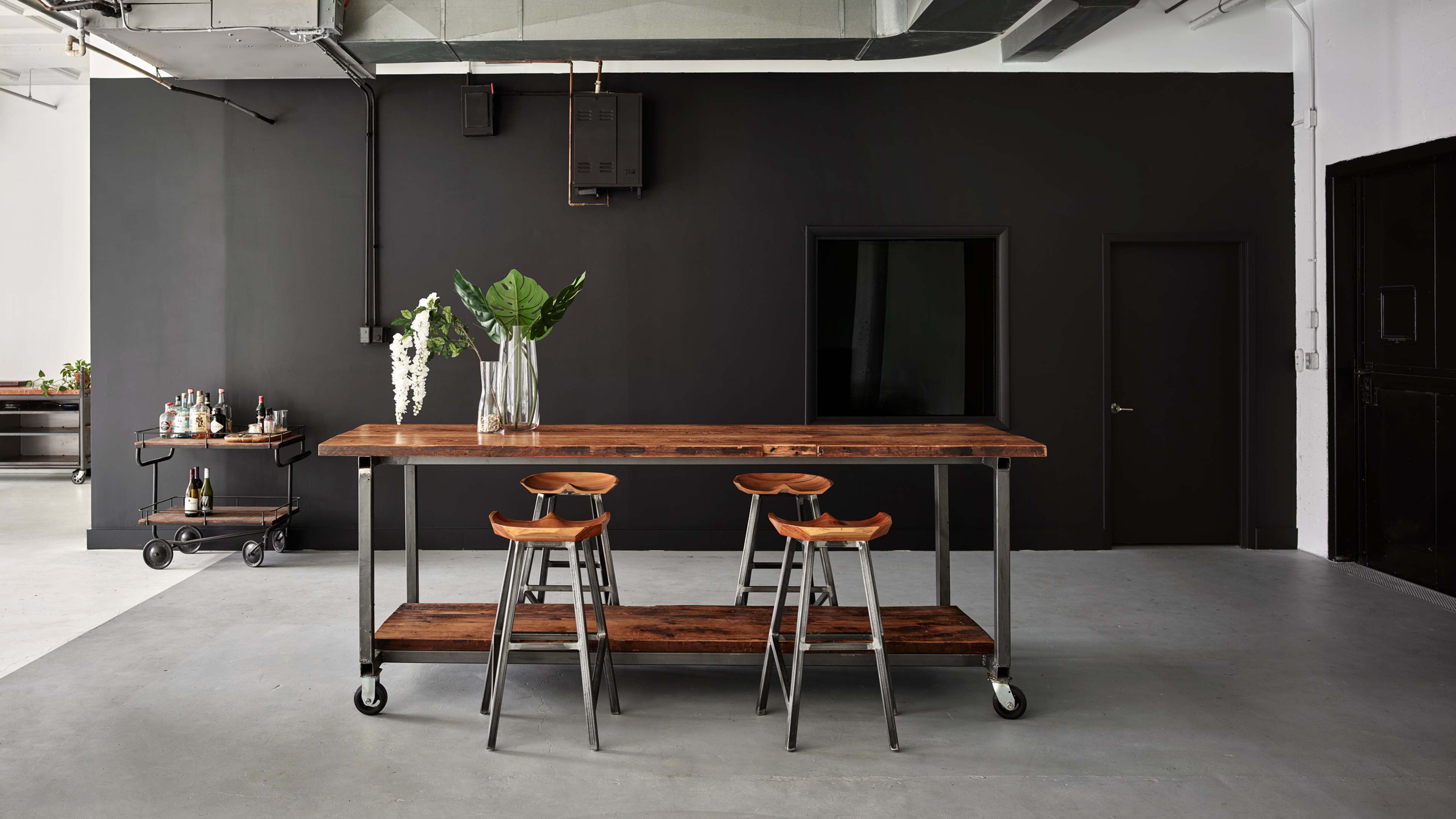 A wooden table with metal legs and three stools is positioned in a minimalist room featuring a black wall and a cart in the background.