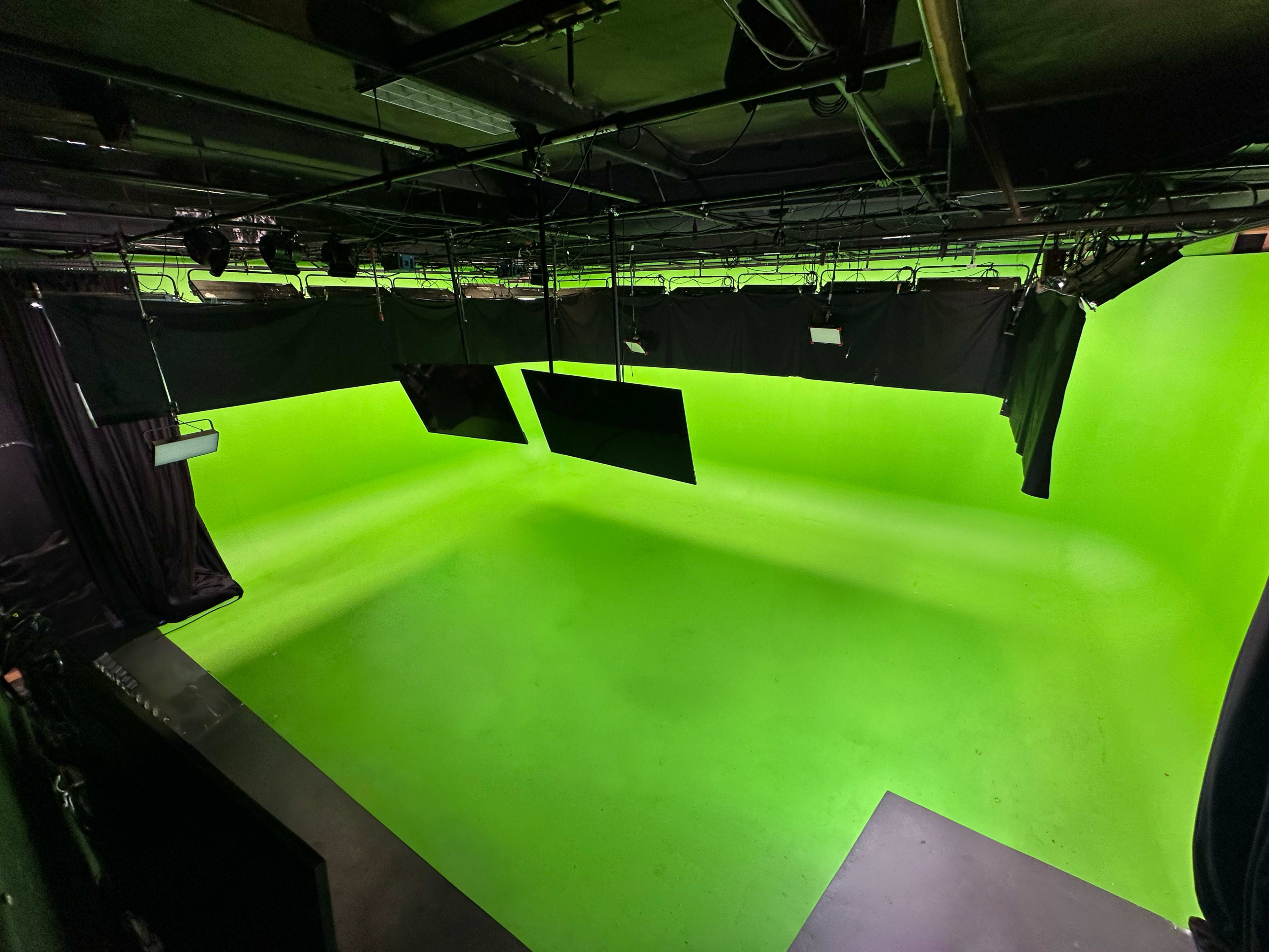 The image shows a green screen studio with a bright green backdrop and overhead lighting fixtures.
