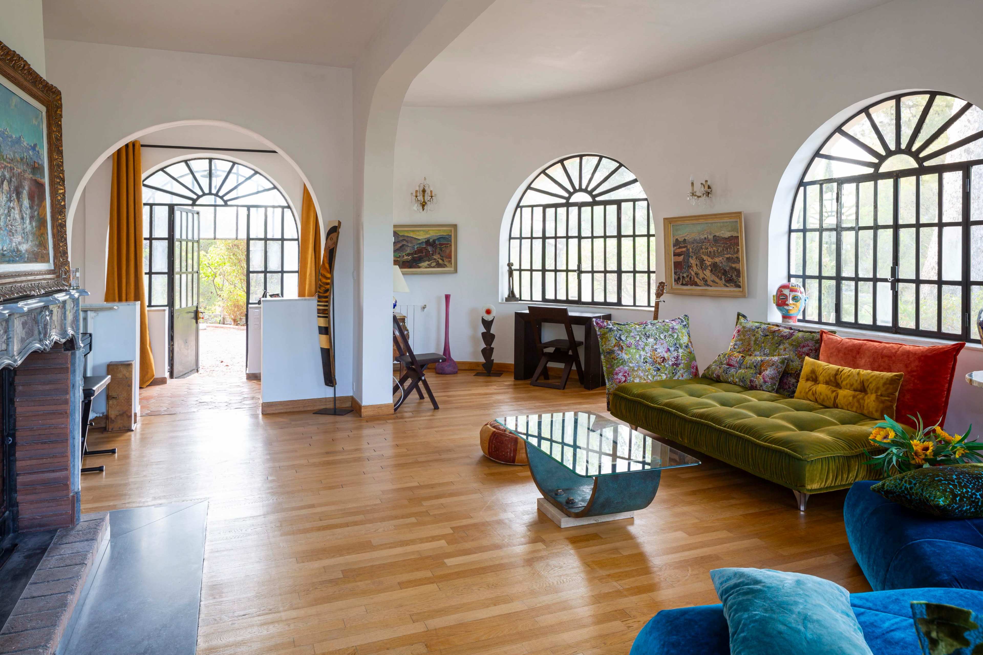 The image shows a bright, open living space with large arched windows, a green velvet sofa, a wooden coffee table, and various artworks on the walls.