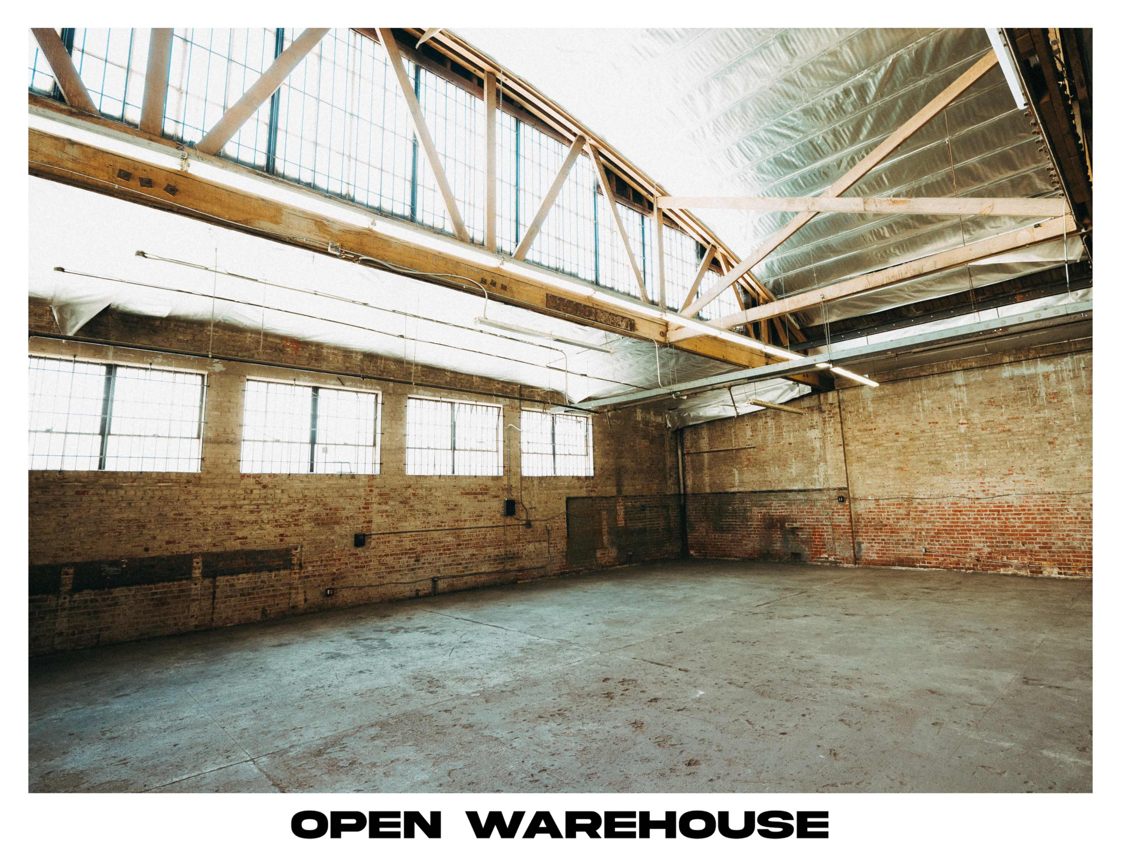 The image shows a large, empty warehouse with high ceilings and large windows, featuring brick walls and a concrete floor.
