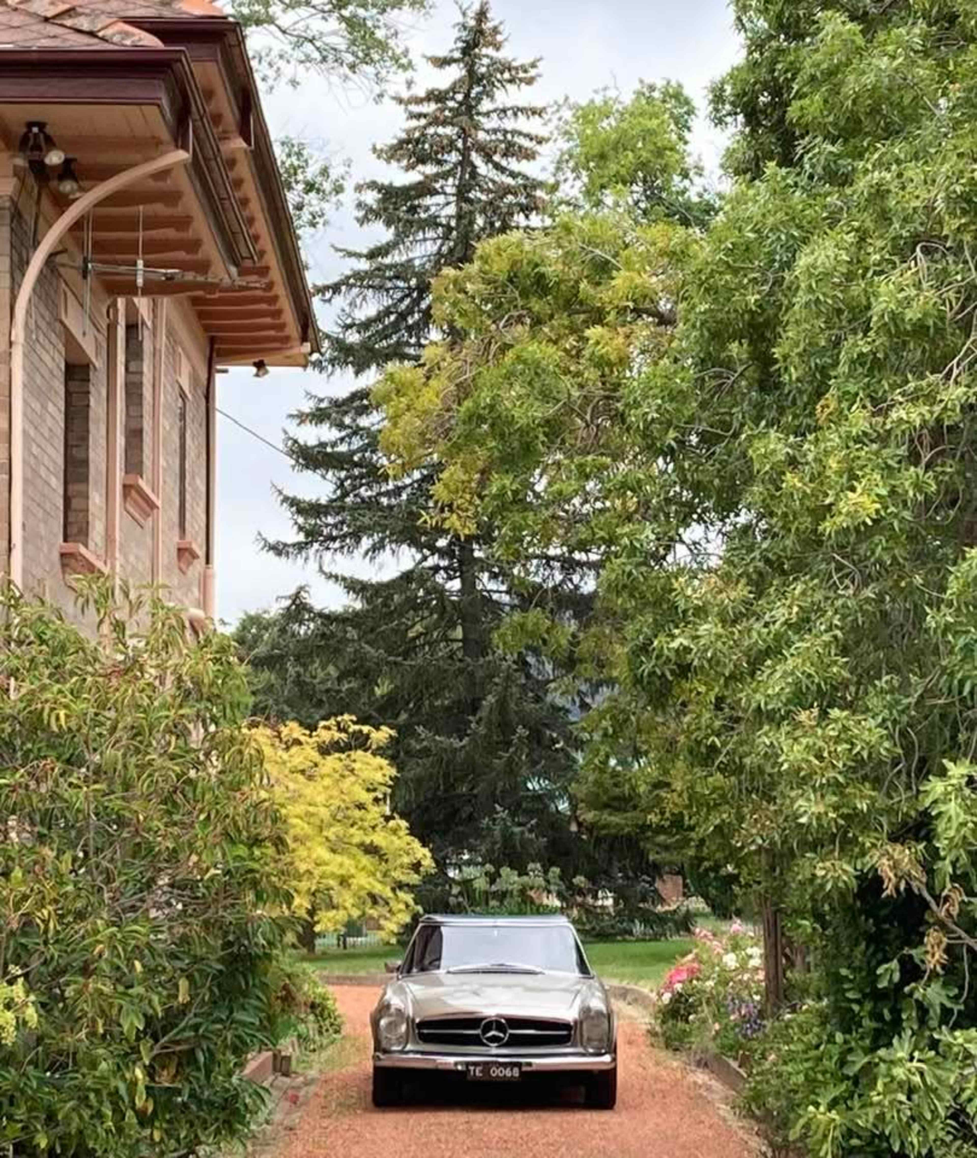 A vintage car is parked on a gravel driveway lined with trees, leading to a large house.