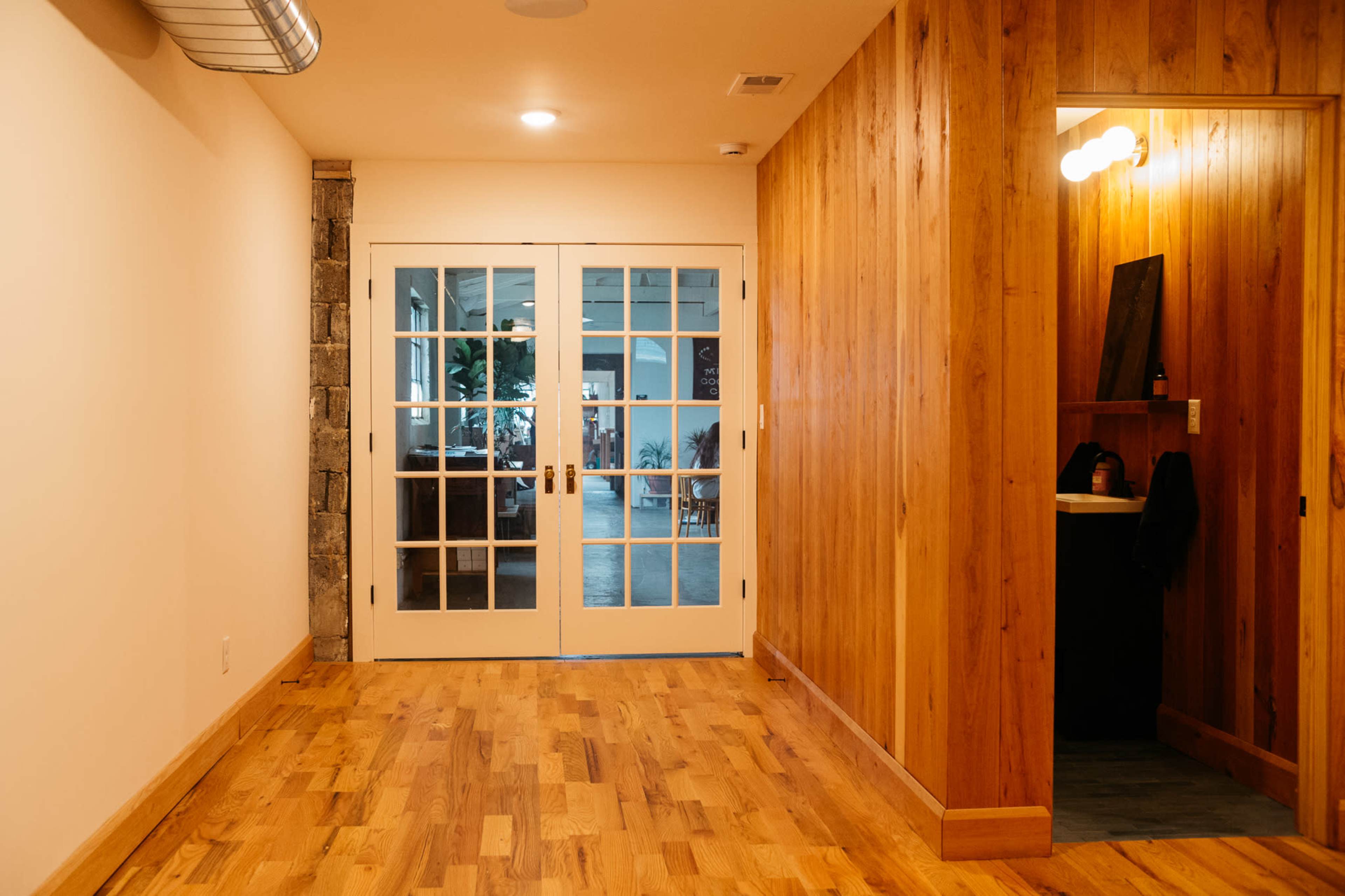 A hallway with wooden flooring leading to double glass doors and an adjacent wooden-paneled area.