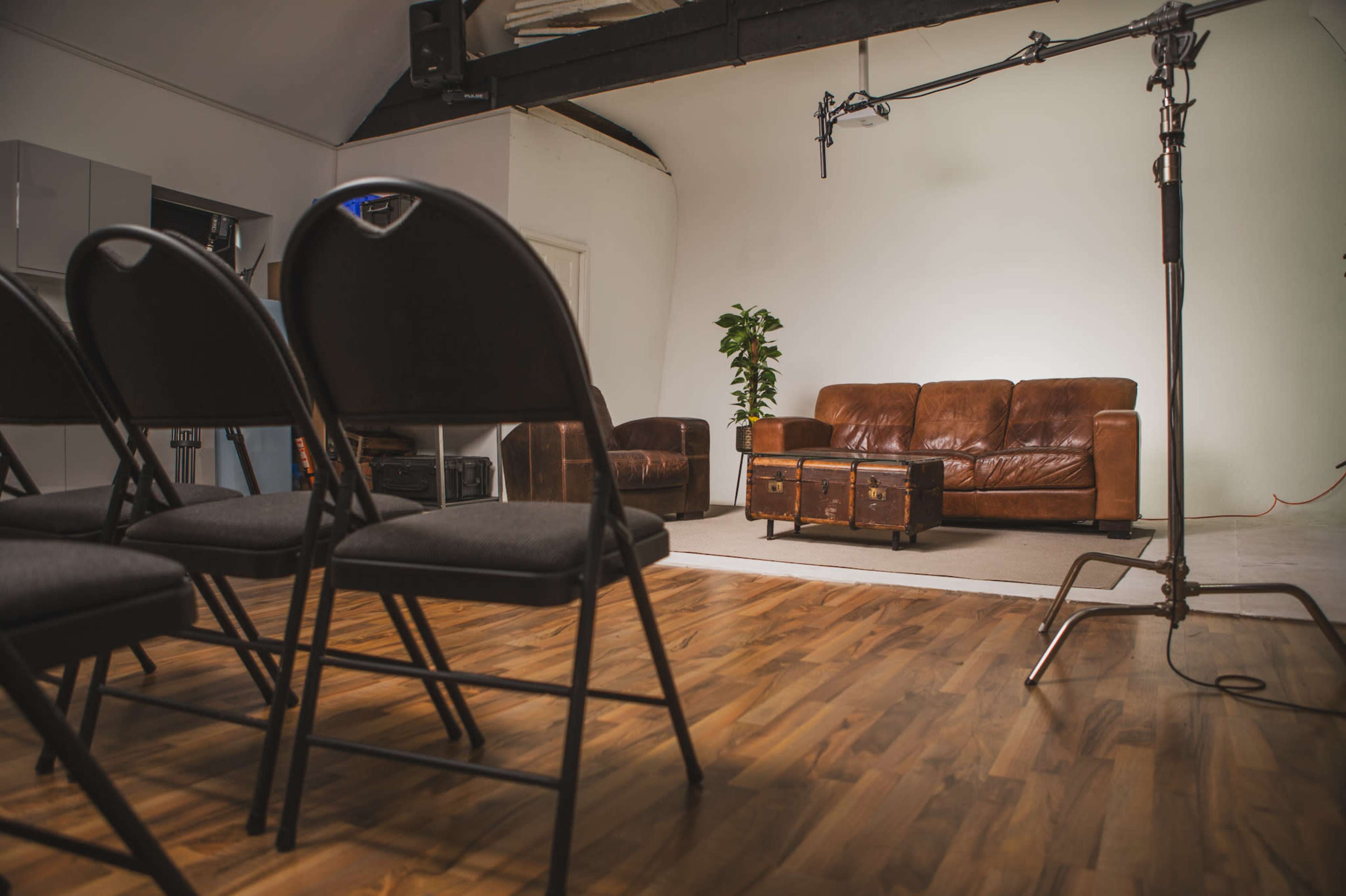 A room with a wooden floor, featuring rows of black folding chairs facing a brown leather sofa and a coffee table, with a microphone setup in front.