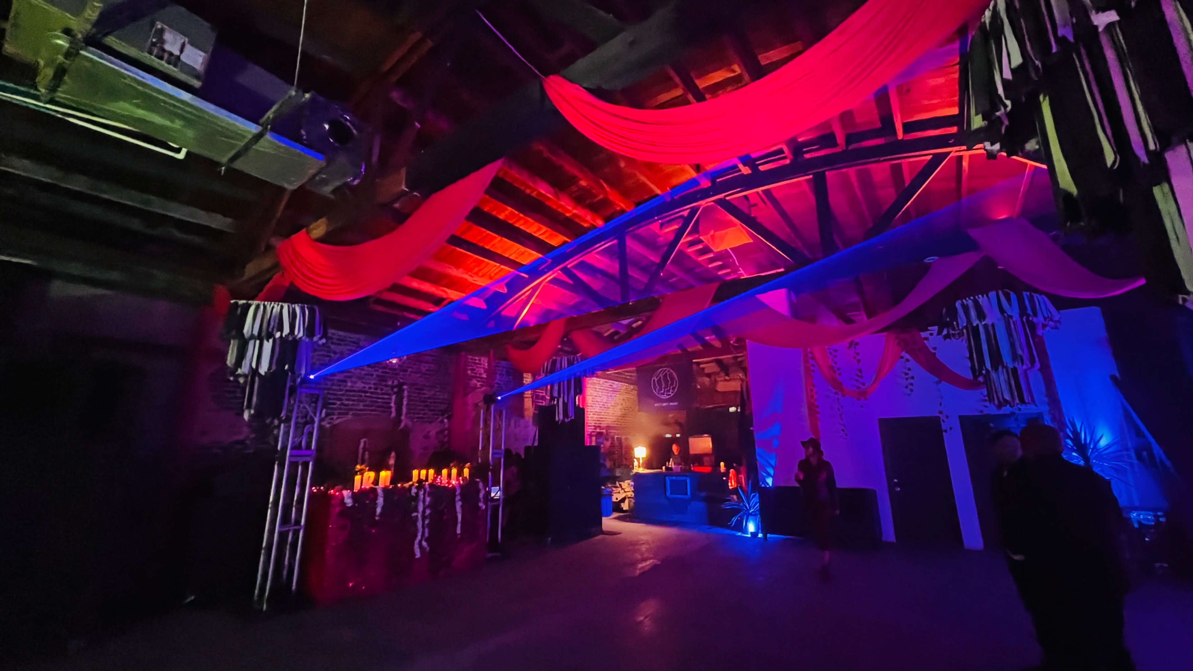 The image shows a dimly lit event space with red and blue drapery, soft lighting, and decorative elements, along with a DJ setup in the background.