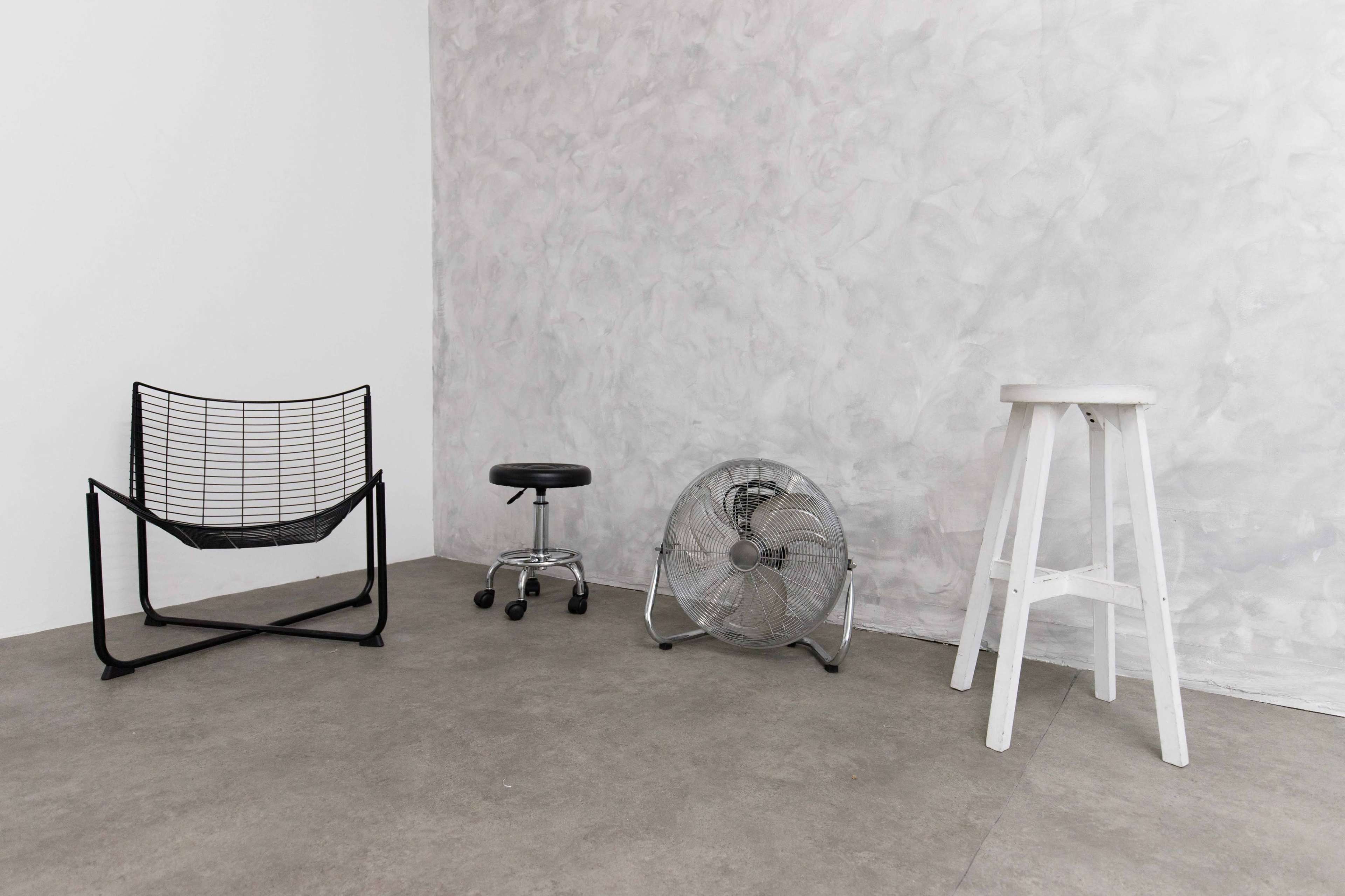 The image shows a minimalist room with a black wire chair, a rolling stool, a metal fan, and a white stool, all situated on a concrete floor against a textured gray wall.