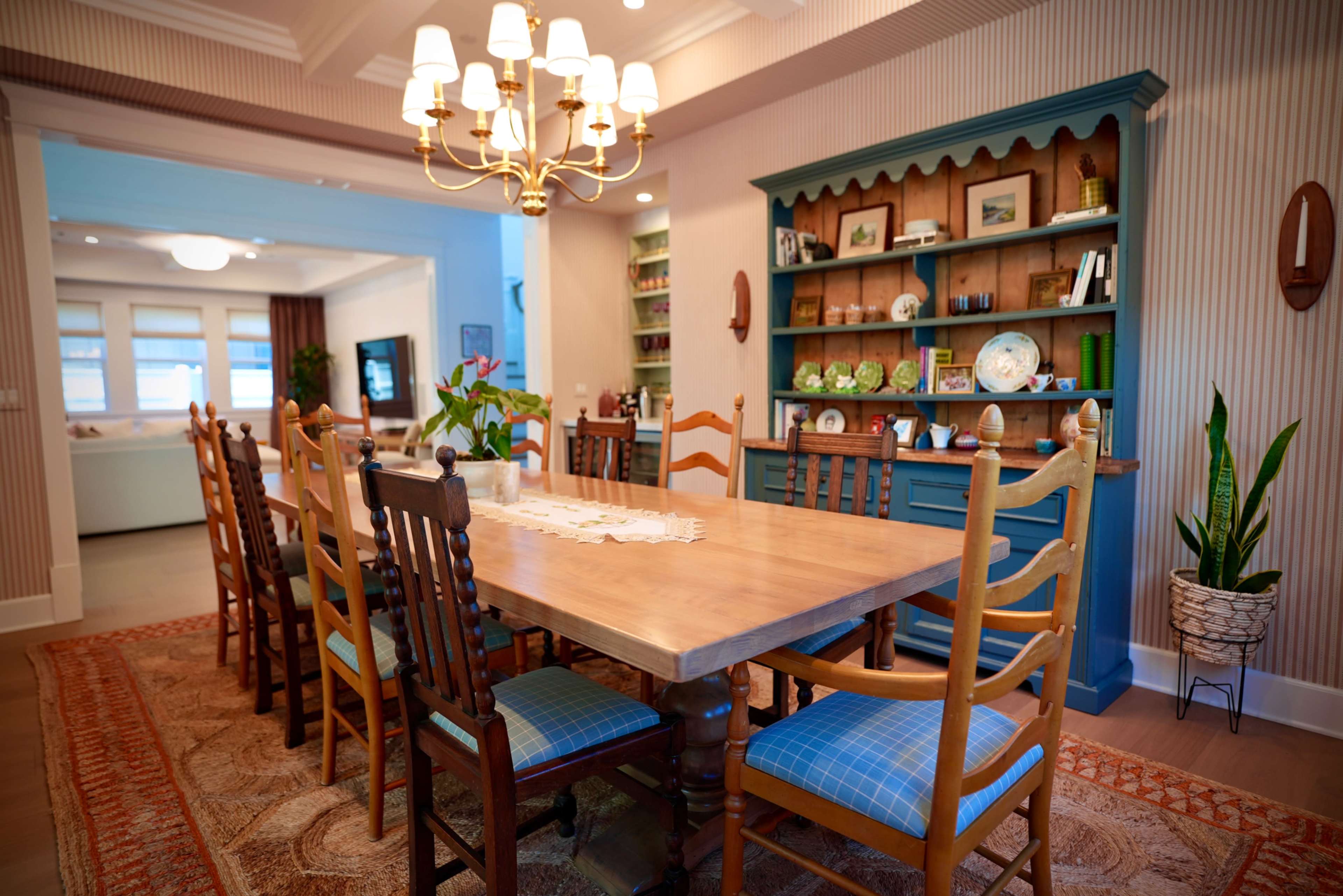 A wooden dining table surrounded by several chairs is set in a brightly lit room with a chandelier, leading to a kitchenette area visible in the background.