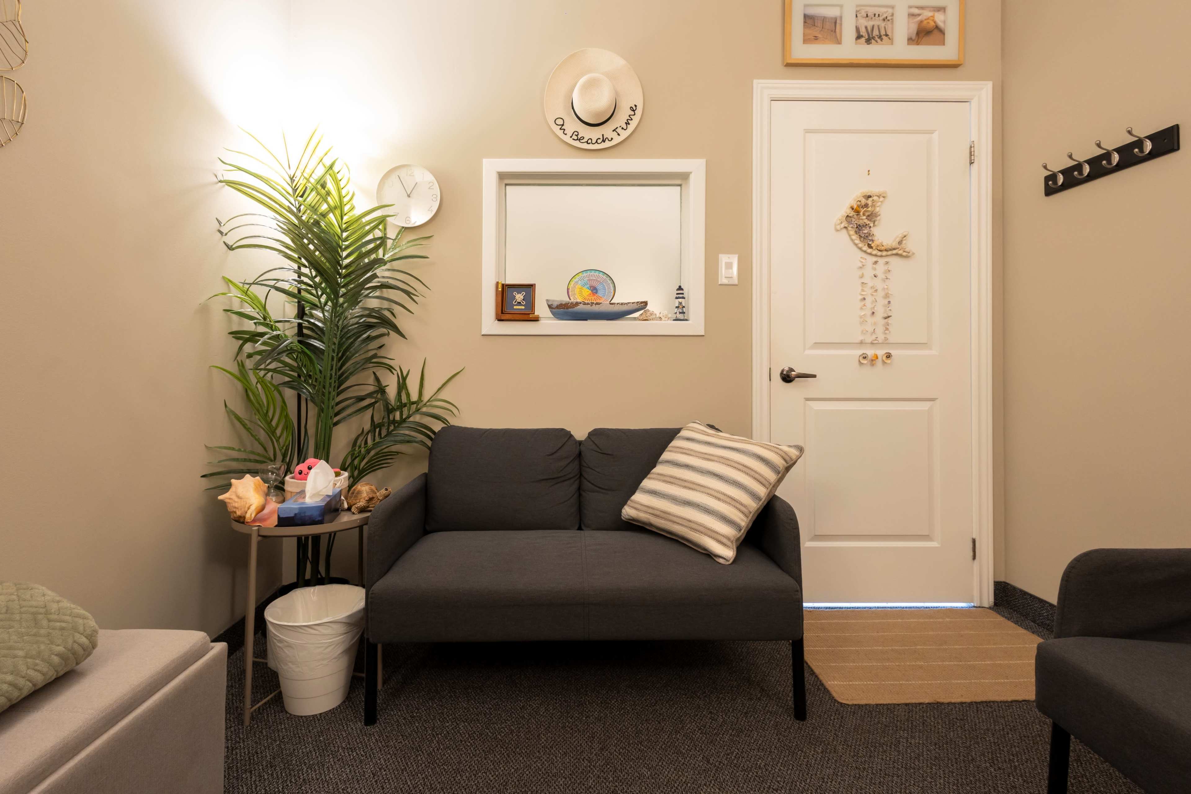 The image shows a small, cozy waiting area with a dark gray sofa, decorative items on the wall, a potted plant, and a door leading to another room.