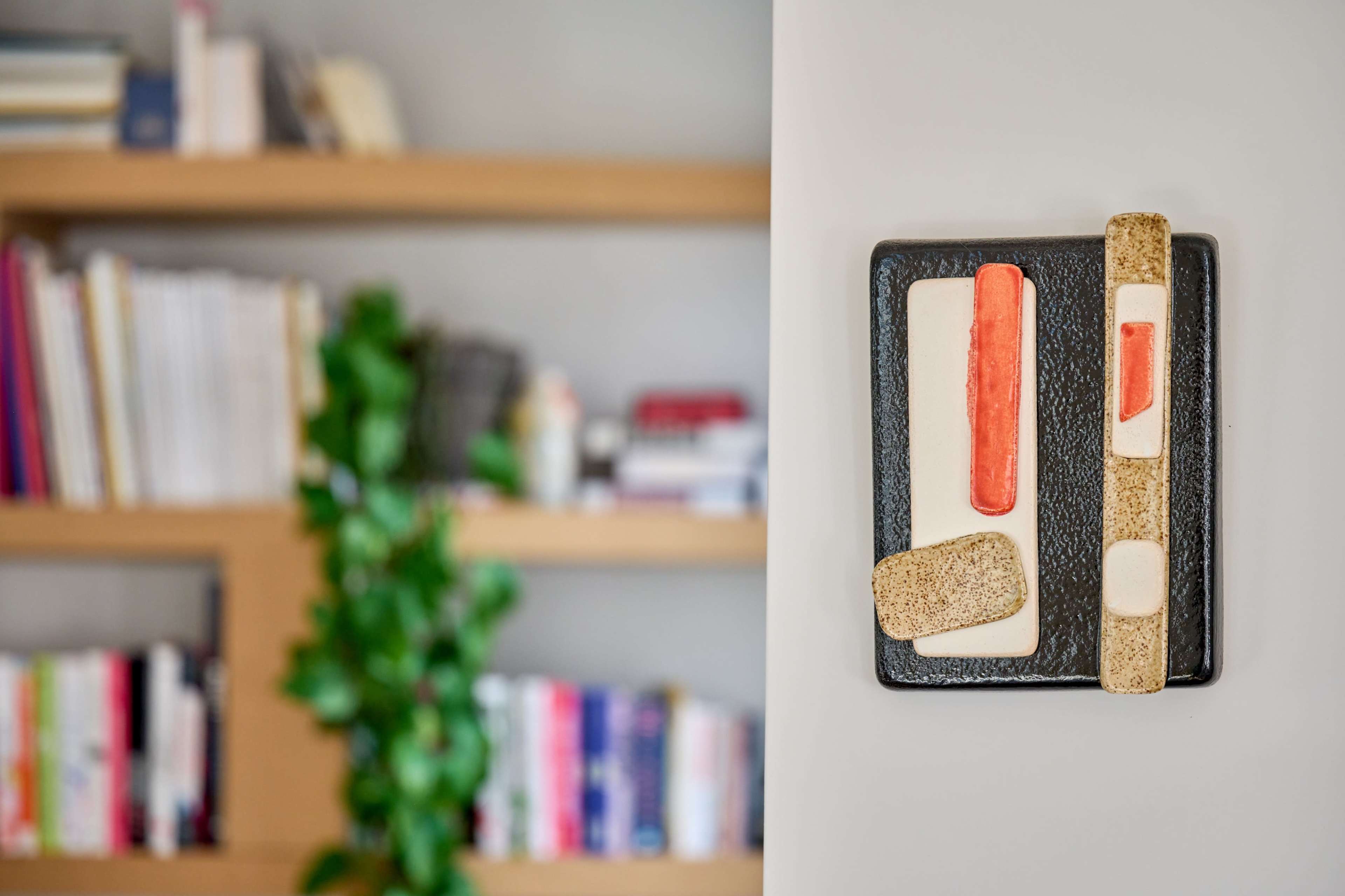 A ceramic artwork mounted on a wall, with a bookshelf filled with books and a plant in the background.