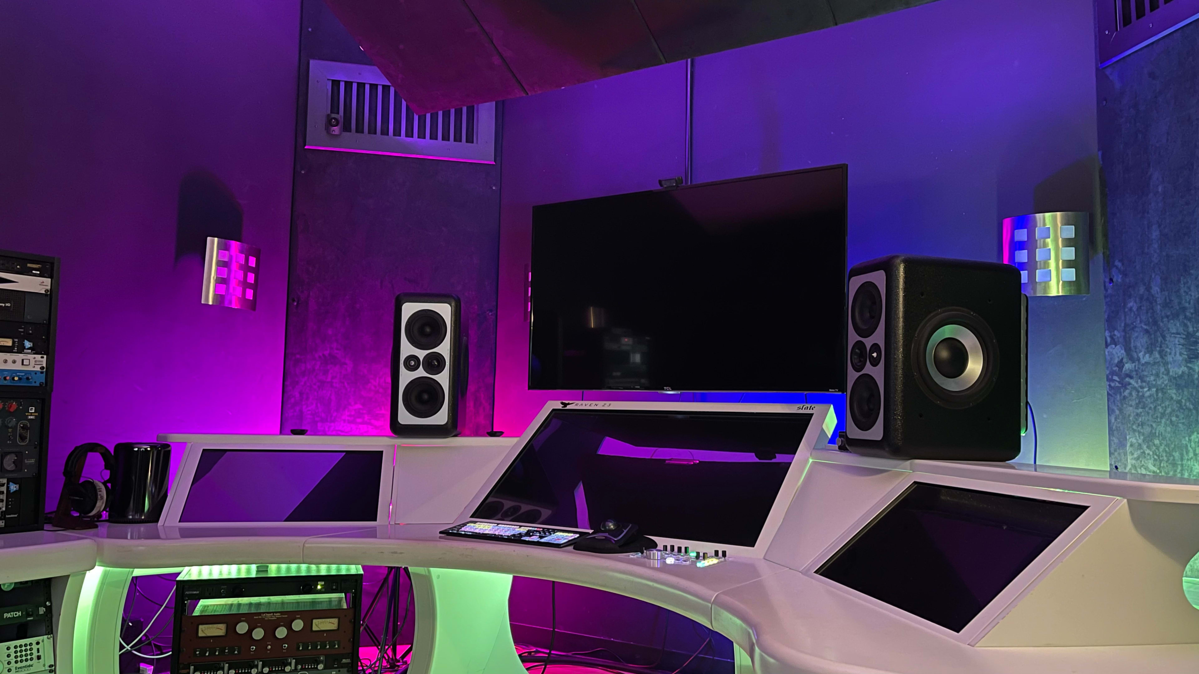 The image shows a modern audio studio with a curved desk, speakers, and a television screen surrounded by colorful ambient lighting.