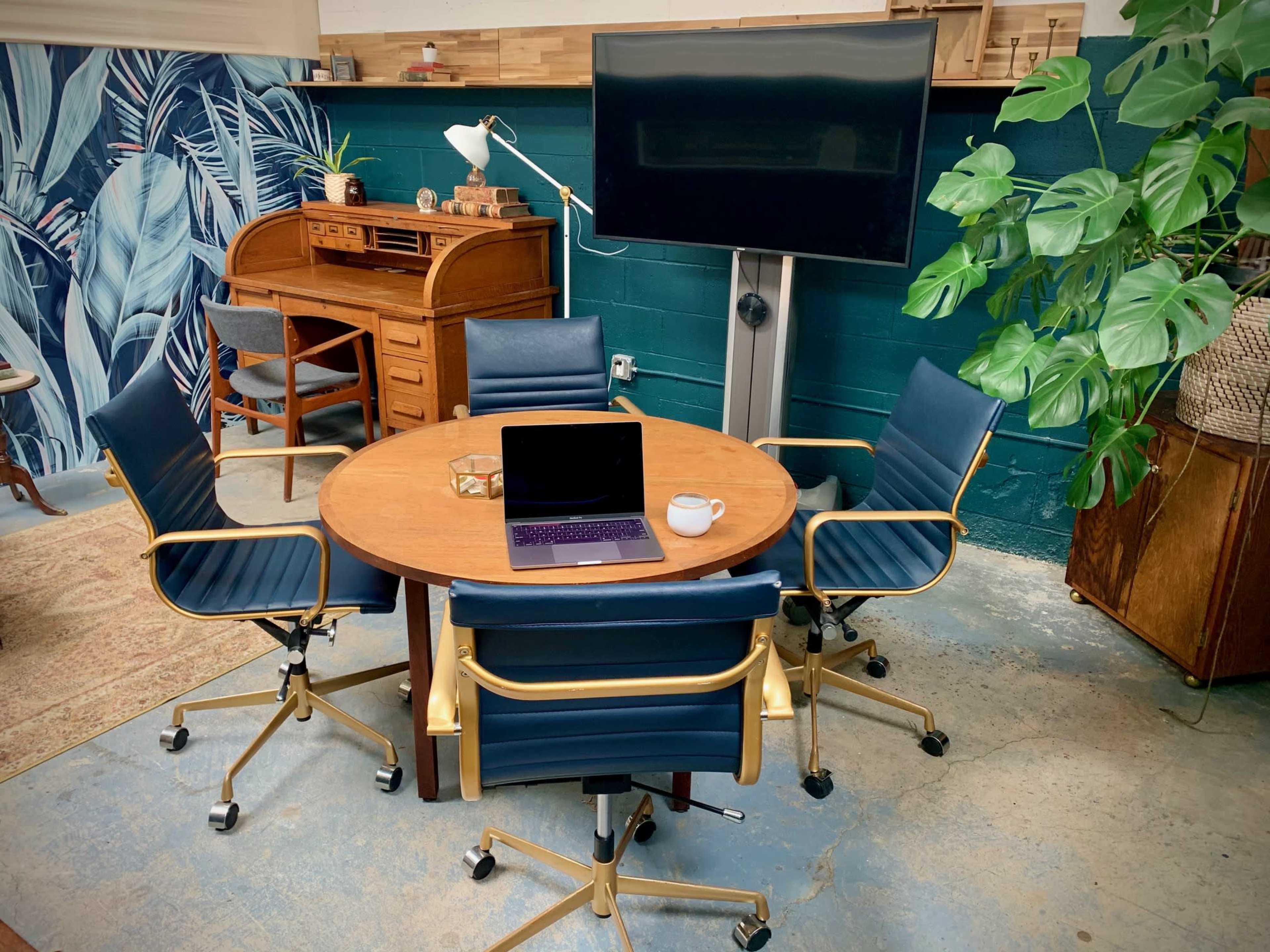 A round wooden table is surrounded by four blue office chairs in a workspace featuring a television and plants.