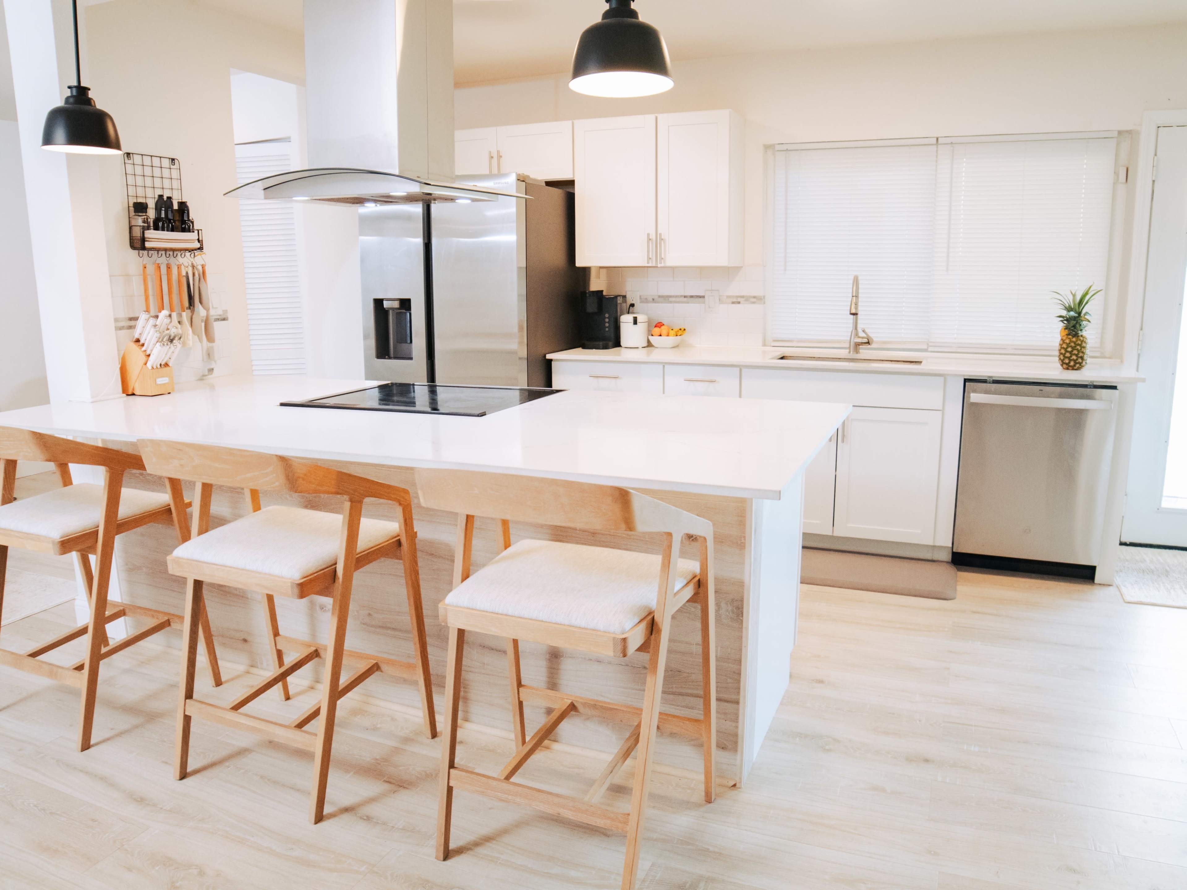 The image shows a modern kitchen with white cabinetry, a central island with four wooden stools, and stainless steel appliances.