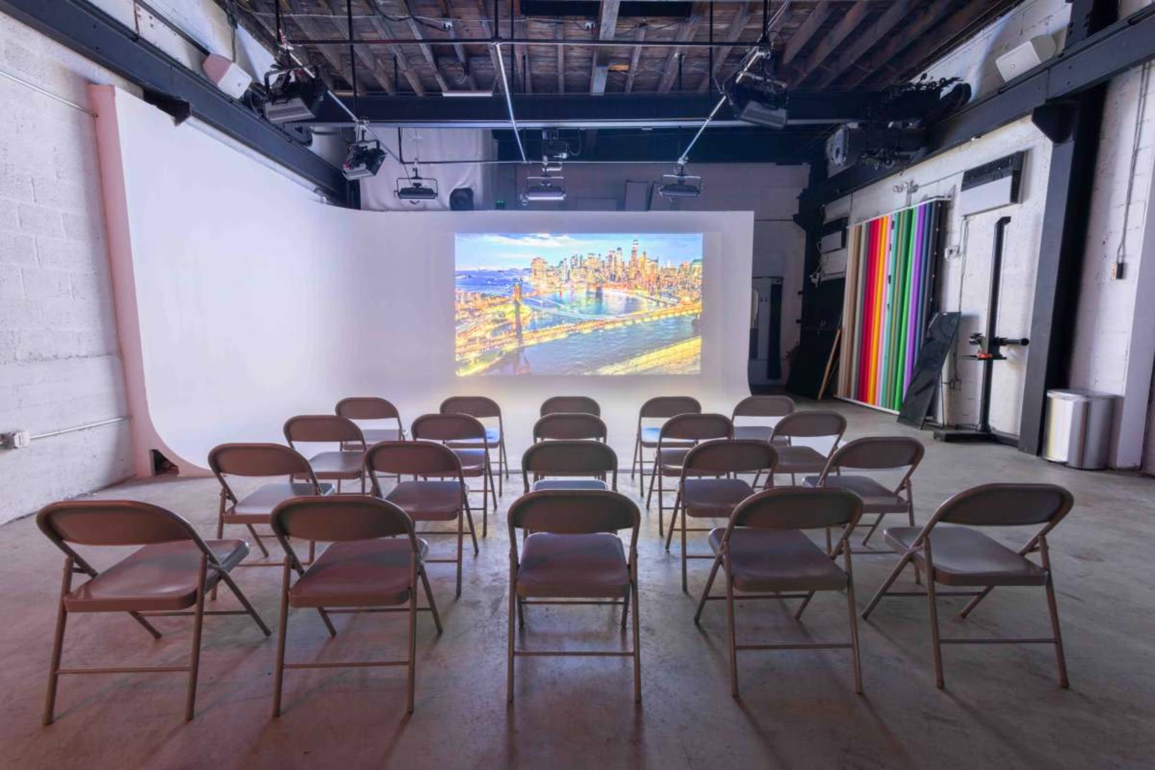 A spacious room with white walls and a projection screen displaying a cityscape, arranged with rows of folding chairs.
