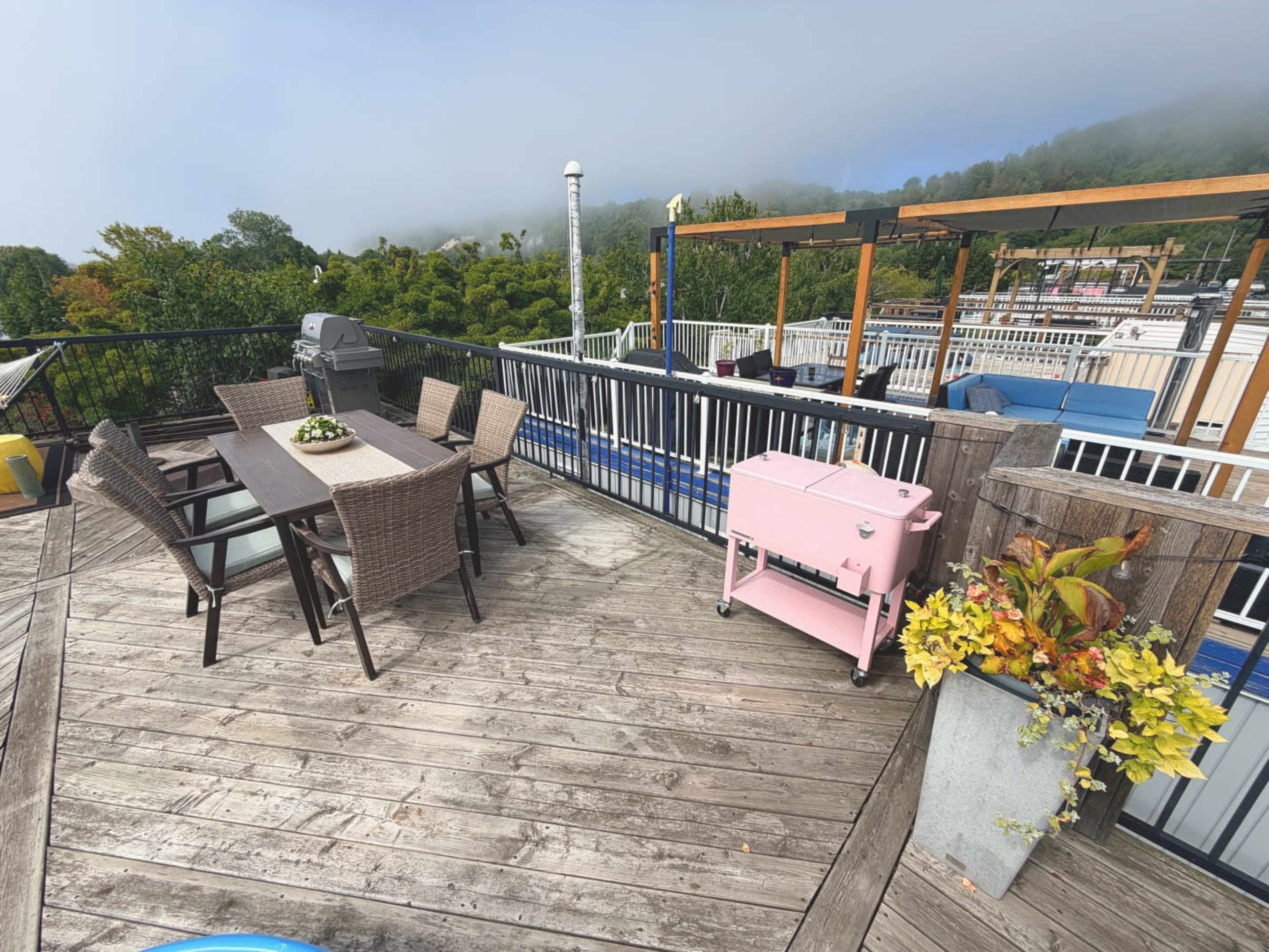 The image shows a rooftop patio featuring a wooden deck with a dining table and chairs, a pink cooler, and lush greenery in the background.