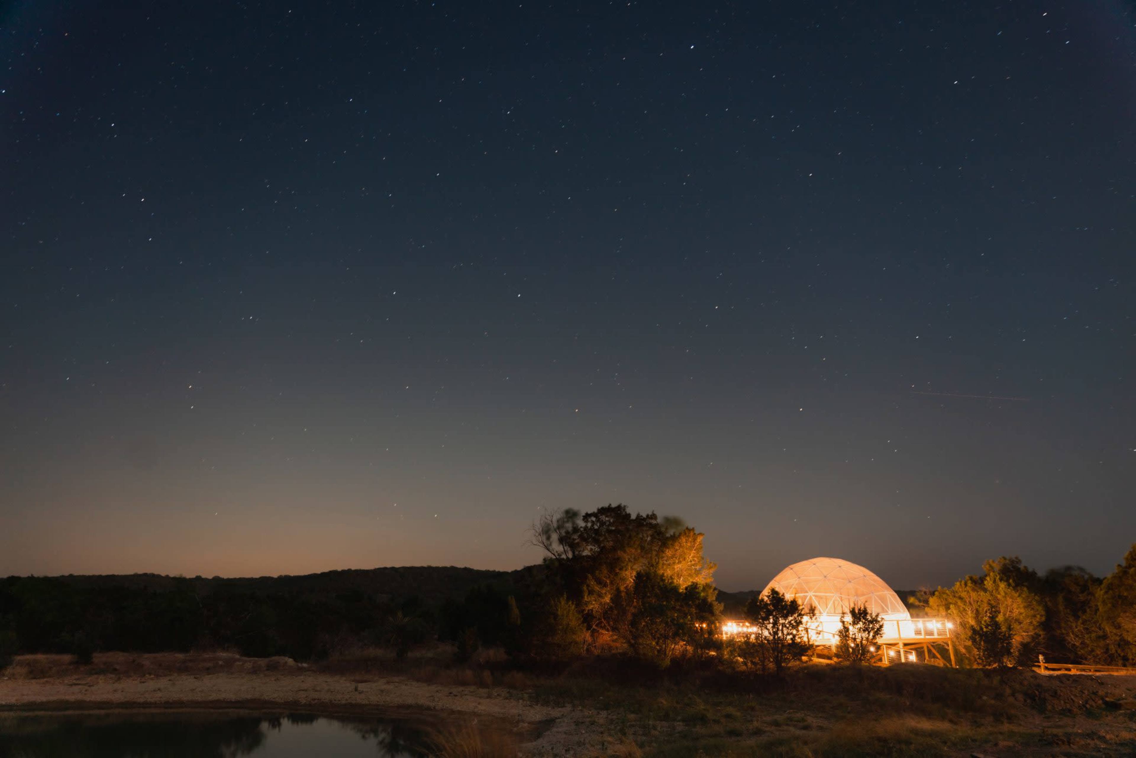 A dome-shaped structure is illuminated at night near a calm body of water, under a starry sky.